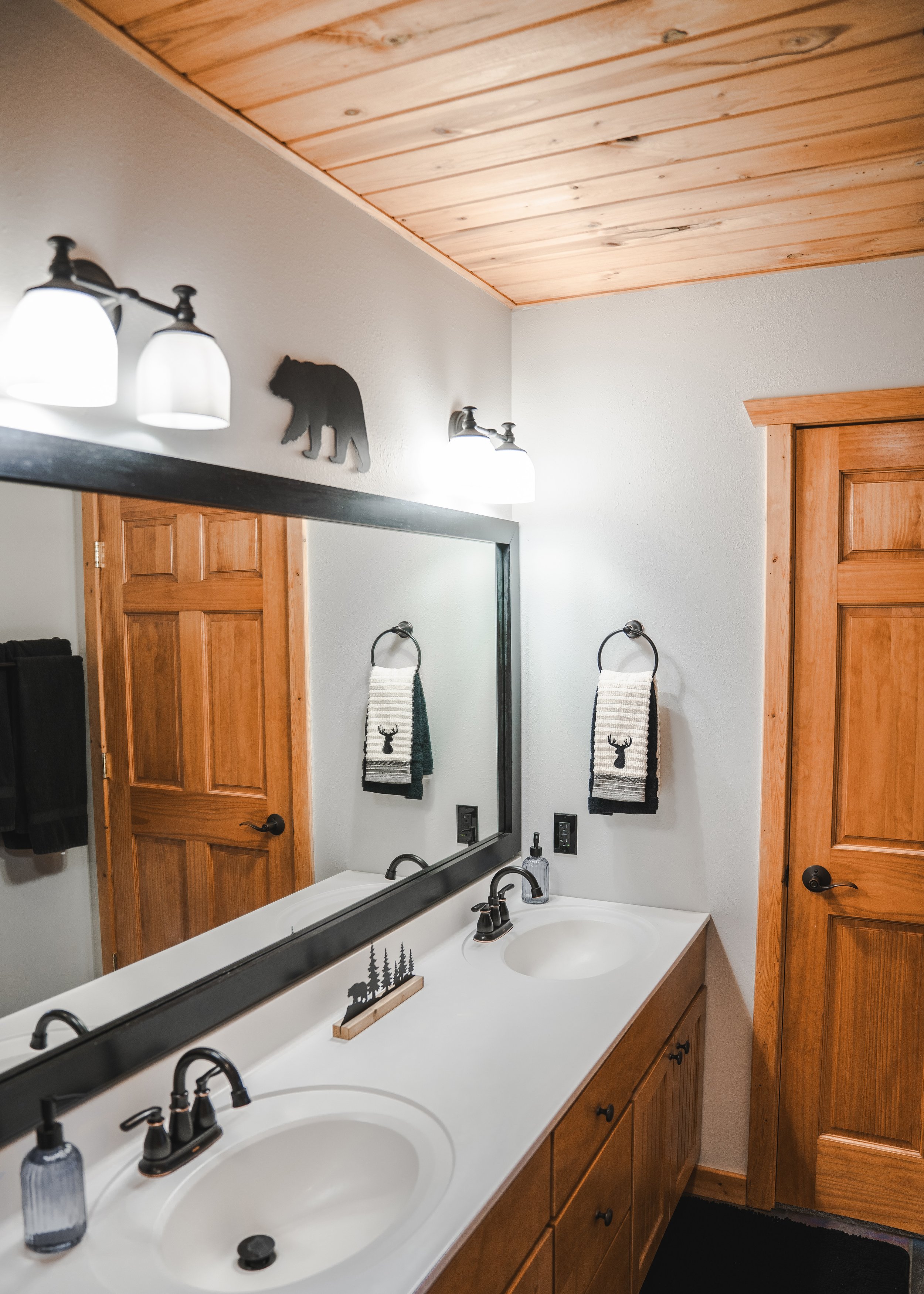 Bathroom with double vanity, wood cabinets, large mirror, wall decor featuring a bear silhouette, and towels with deer print, wooden ceiling, and wooden doors.