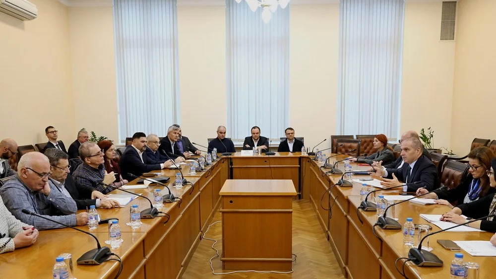 A meeting room with about twenty people seated around a U-shaped conference table, with microphones, water bottles, papers, and laptops, in a well-lit room with large vertical blinds and beige walls.