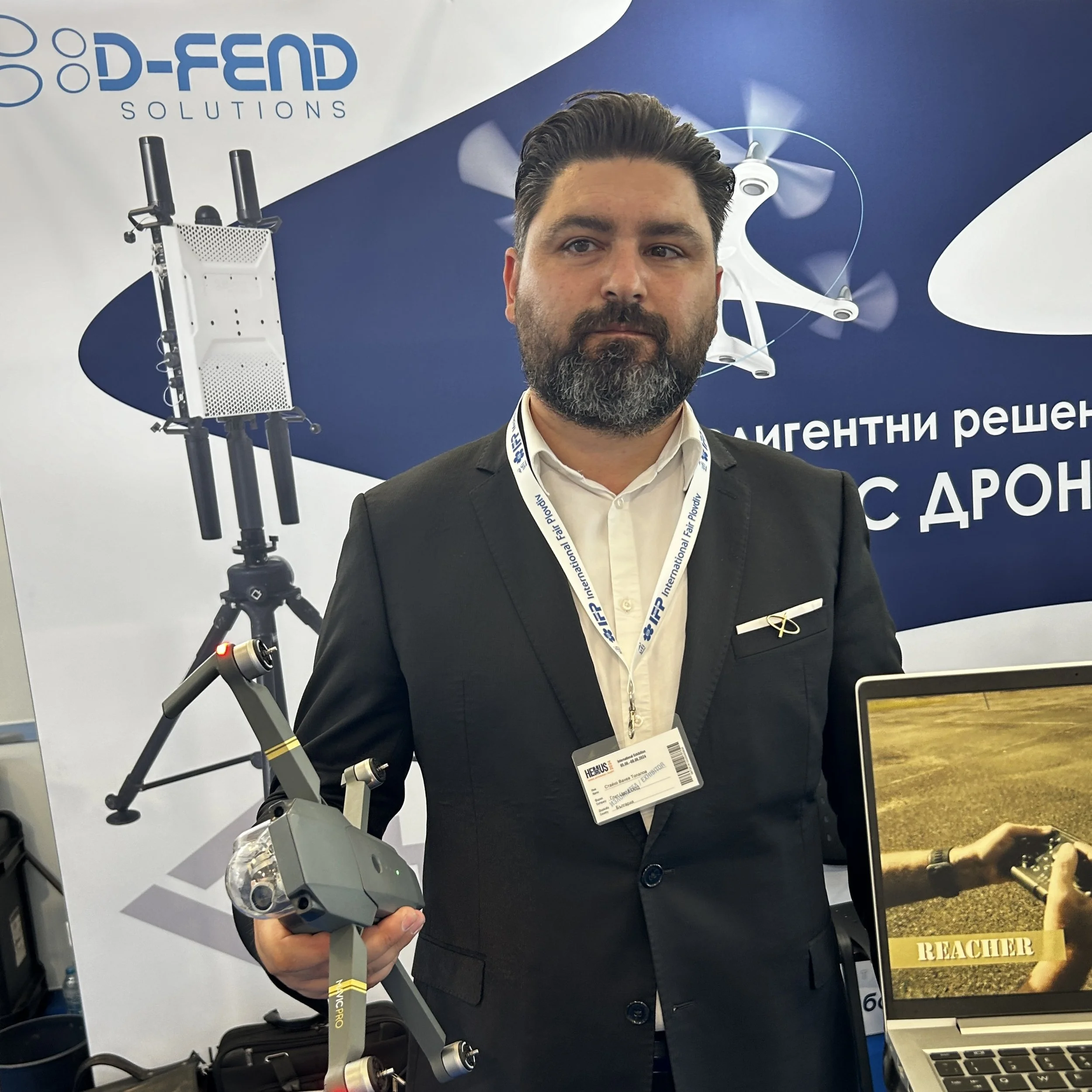 A man in a suit holding a drone at a technology exhibition with a banner in the background displaying drone solutions and a laptop showing an image of someone holding a drone remote control.