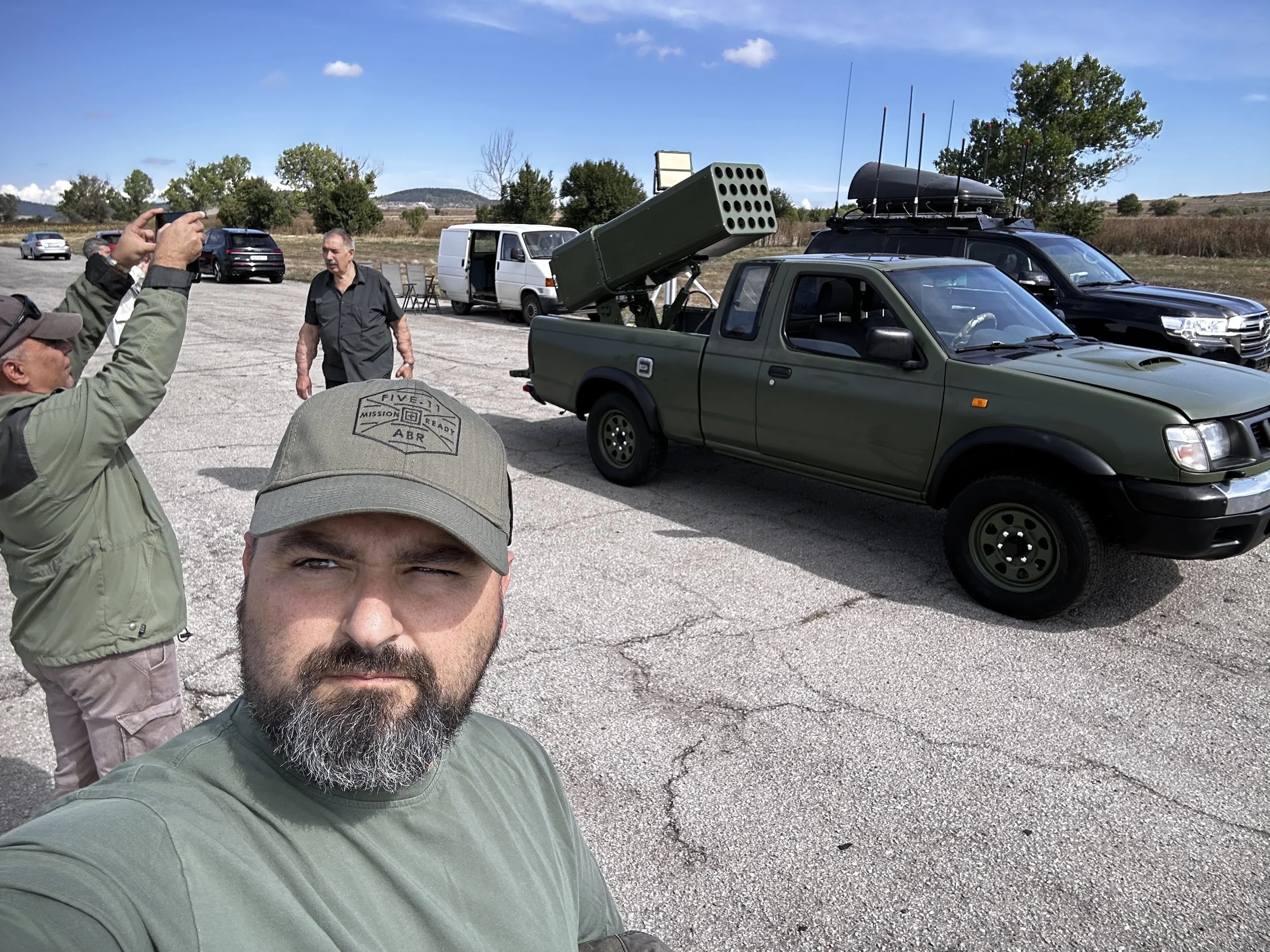 Group of men outdoors with military vehicles, including a green pickup truck with a mounted missile launcher and a black vehicle with antenna arrays, on a cracked paved surface with trees and blue sky in the background.