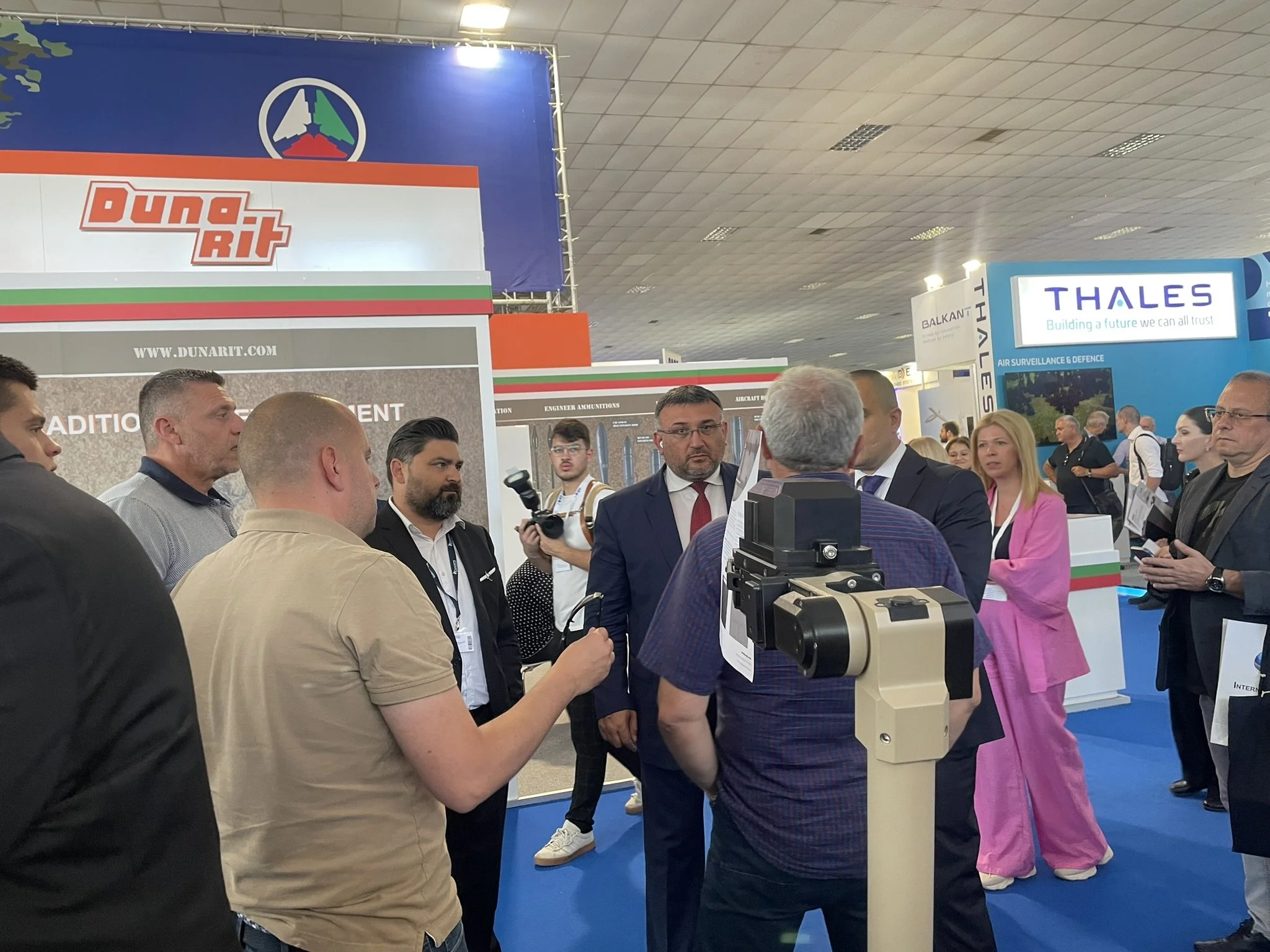 Group of people at a conference or trade show, engaging in conversation, with booths and company signs in the background, including DunaRit and Thales.