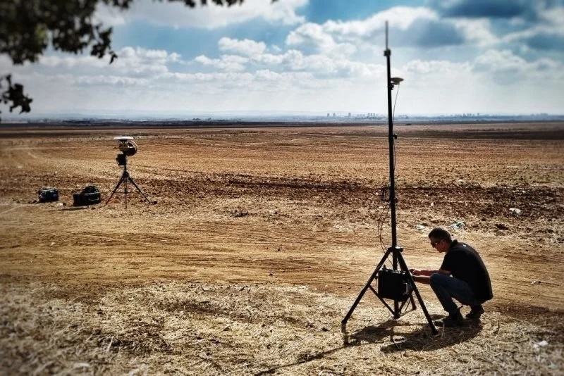 A person setting up camera equipment on a tripod in a vast, dry, open field with a distant city skyline and cloudy sky in the background.