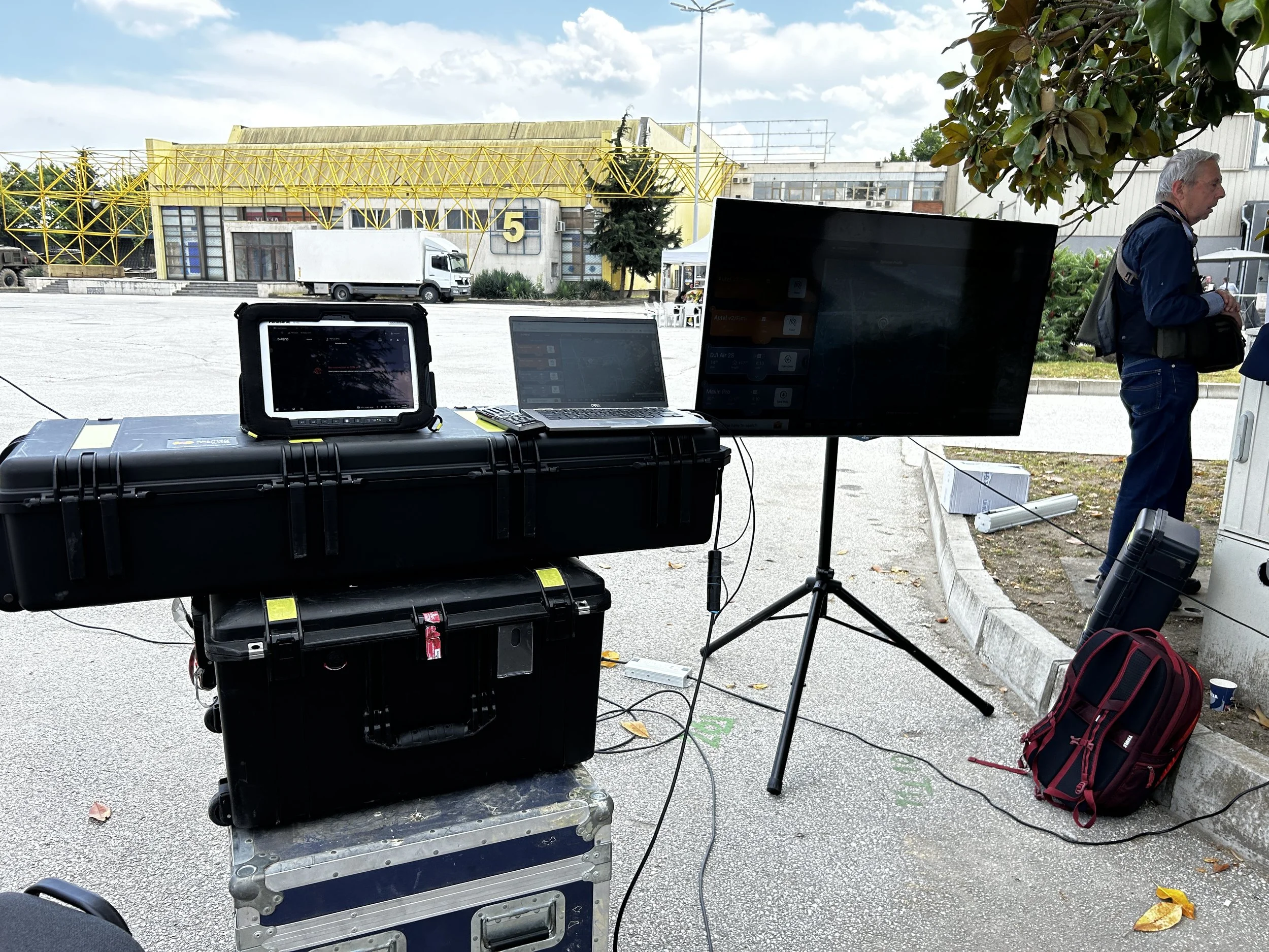 Outdoor scene with electronic equipment including a monitor, laptop, and large screen on tripods, set up on a paved area near a curb, with a person in a dark jacket and jeans standing to the right and Trees and buildings in the background.