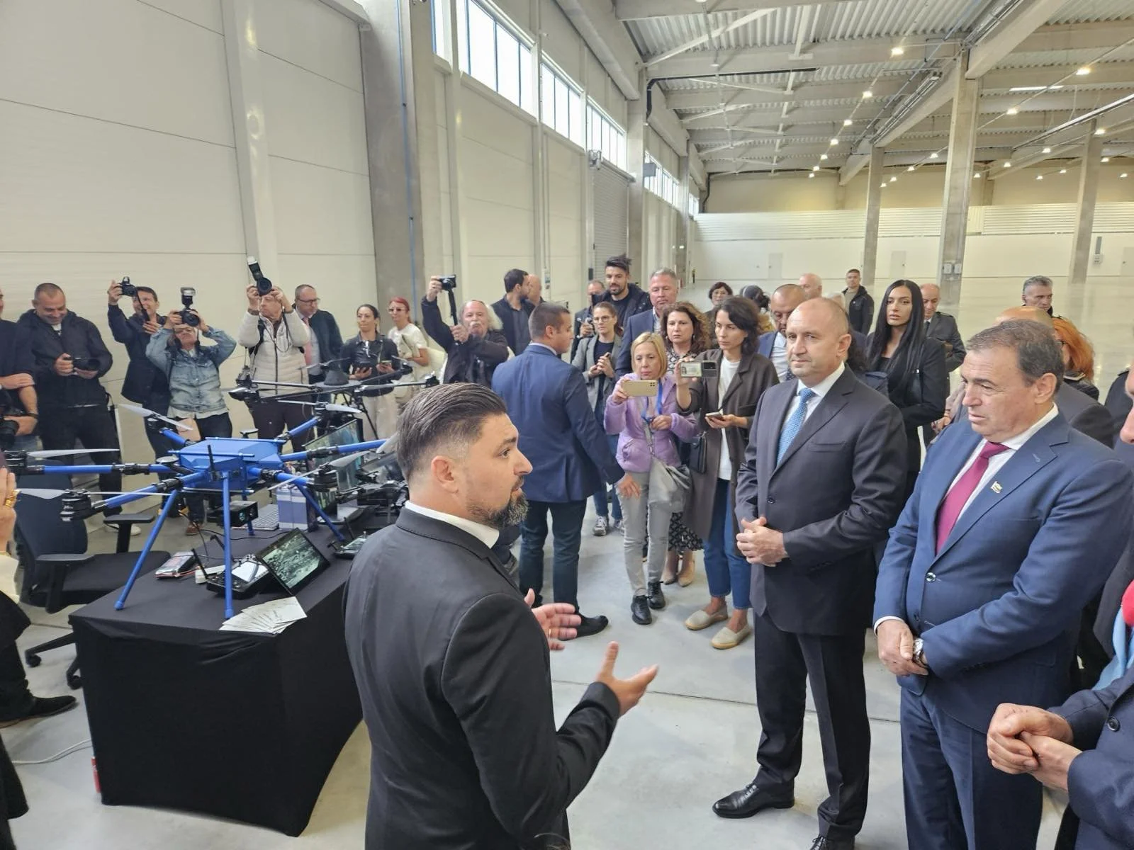 A man in a black suit is speaking to a group of people, including several men in suits, inside a spacious industrial facility with high ceilings. Many people are taking photos or recording videos, and there is a drone on a table nearby.