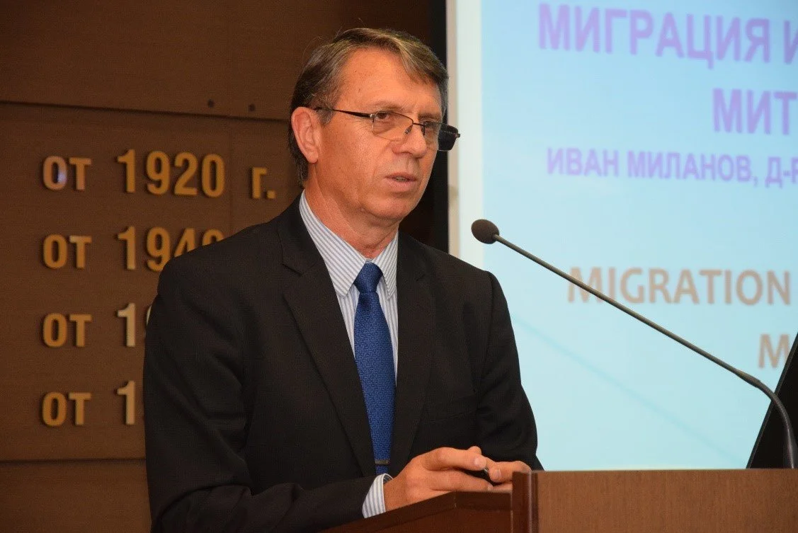 A man in a suit and tie speaking at a podium during a presentation or conference, with a projected slide in the background.