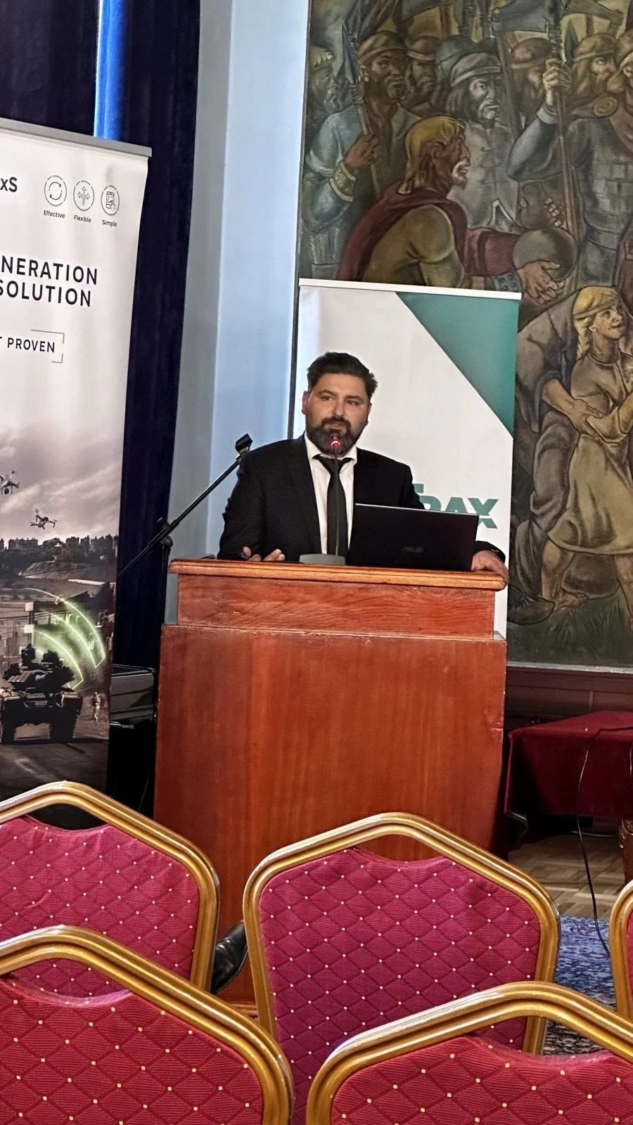 A man with dark hair and a beard, wearing a black suit and tie, stands behind a wooden podium with a microphone, giving a presentation in a conference room. There are red-upholstered chairs with gold trim in the foreground, and two large banners are visible behind him. One banner shows images of drones and a tank with the text 'Generation Solution,' and the other features a logo and text partially visible. The background includes a mural of historical soldiers.