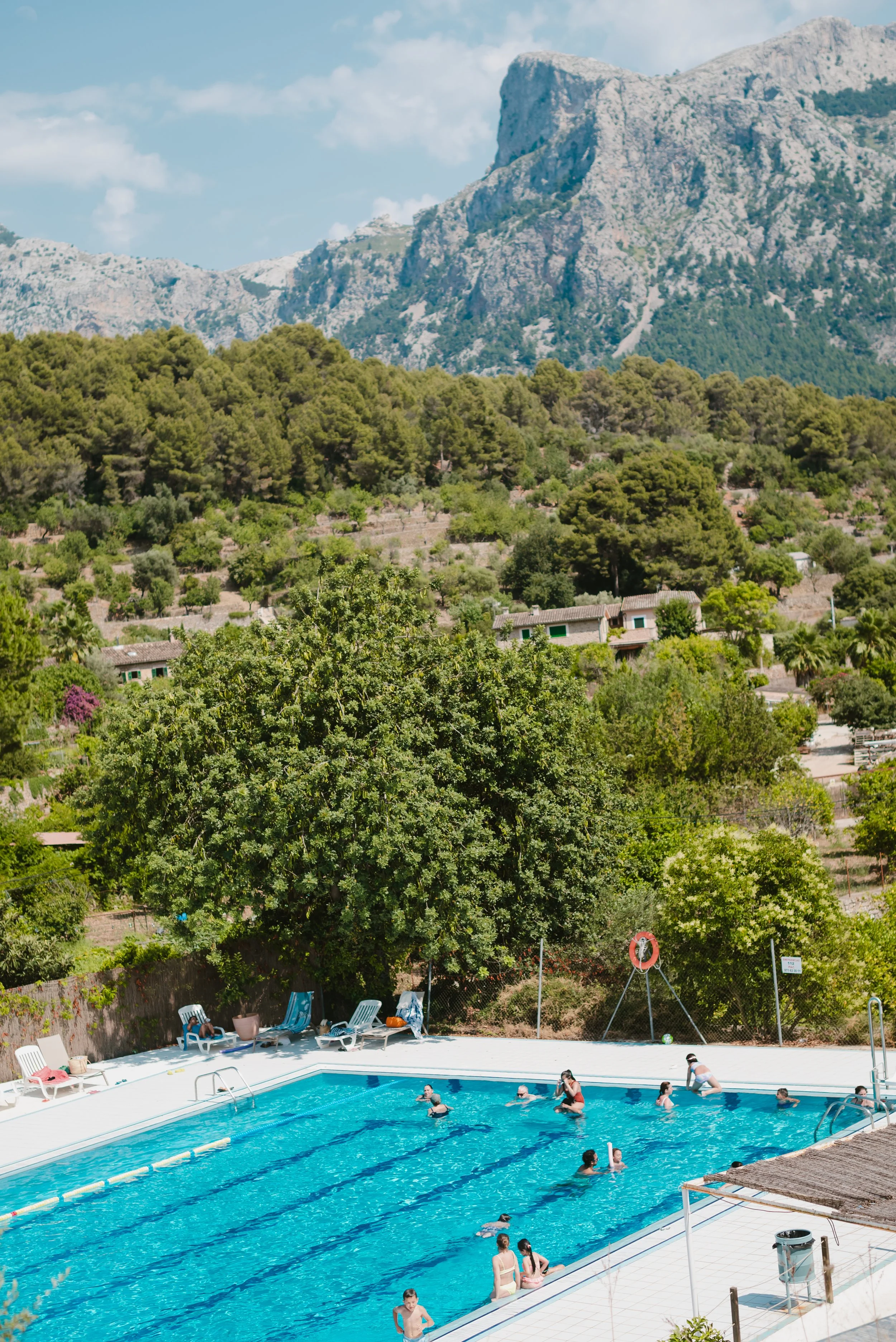 People swimming and relaxing in an outdoor pool with lush green trees and mountainous landscape in the background.