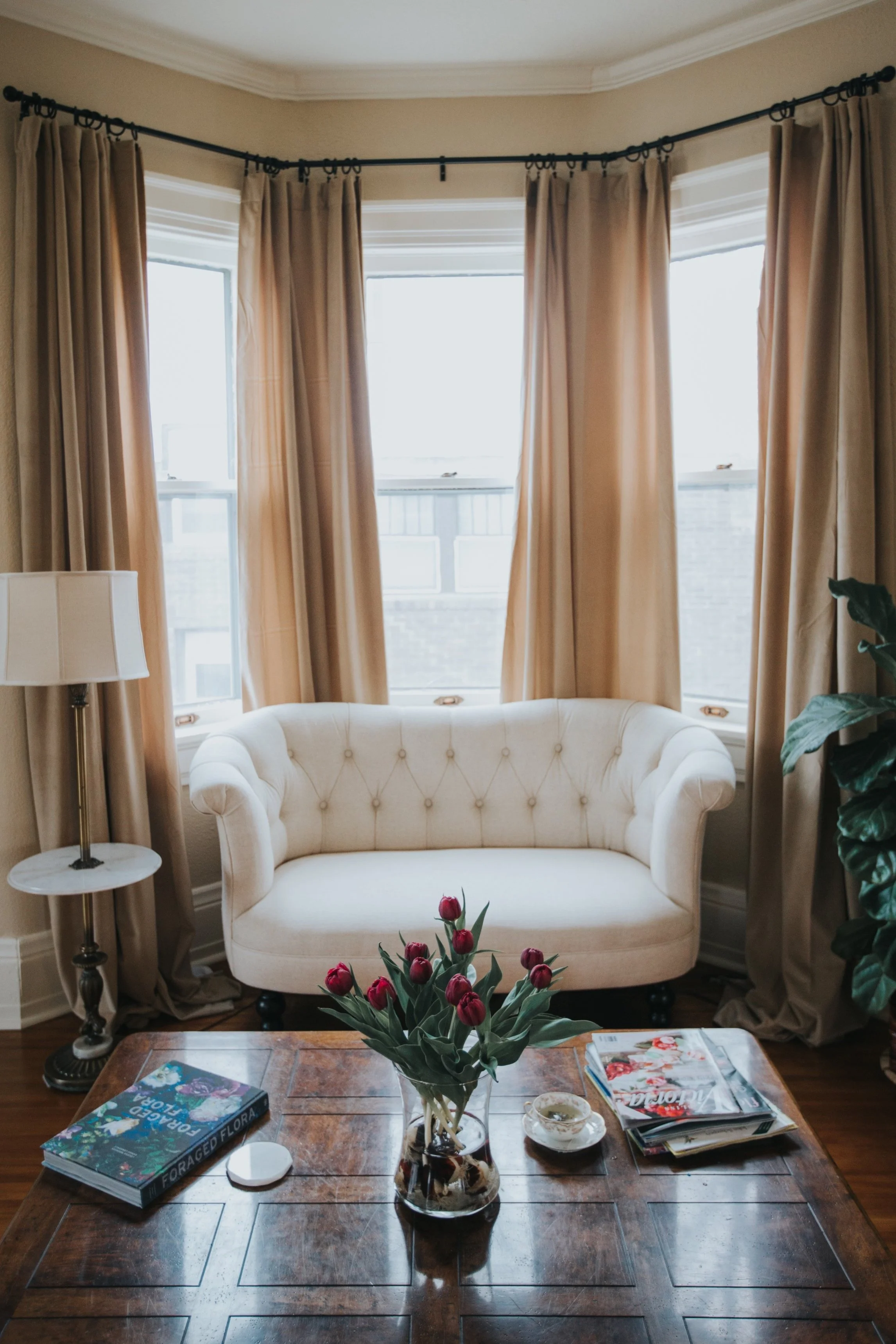 Sunlit traditional living room designed by Summer Glen Design with a cream tufted loveseat, floor-length neutral drapery, antique wood coffee table, and warm natural light.