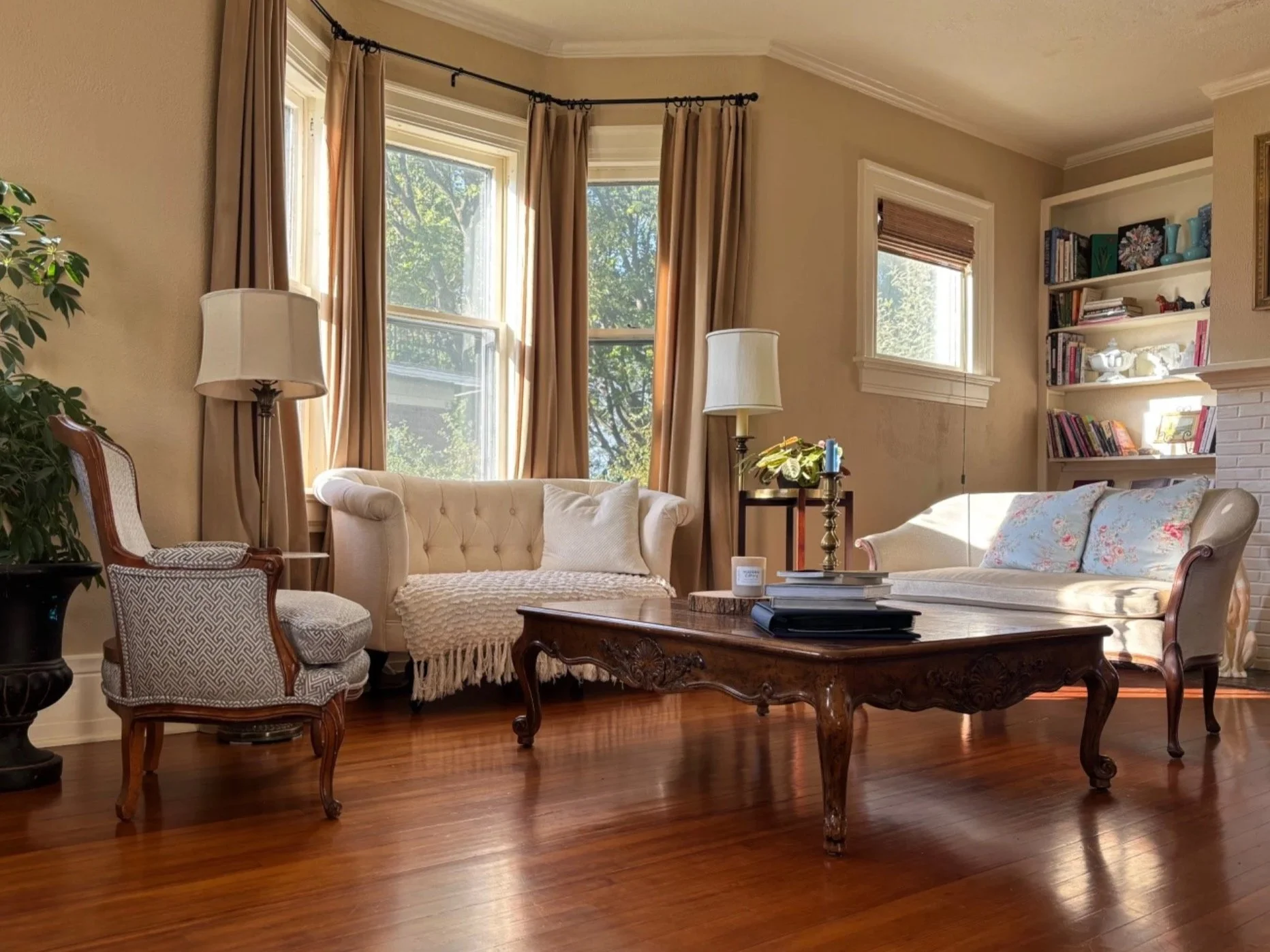Traditional living room designed by Summer Glen Design featuring antique wood furnishings, a cream tufted loveseat, patterned accent chairs, warm hardwood floors, and neutral drapery.