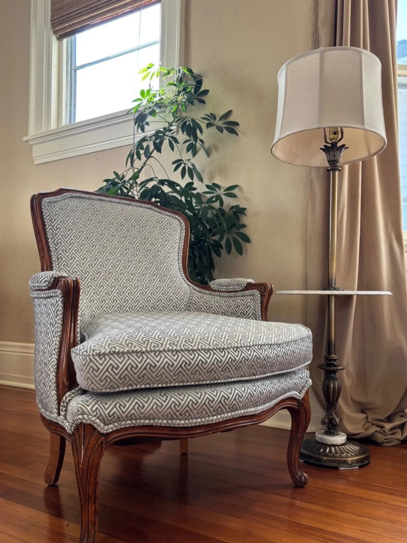 Traditional wood-framed accent chair upholstered in a neutral geometric fabric, paired with a vintage brass floor lamp, greenery, and hardwood floors.