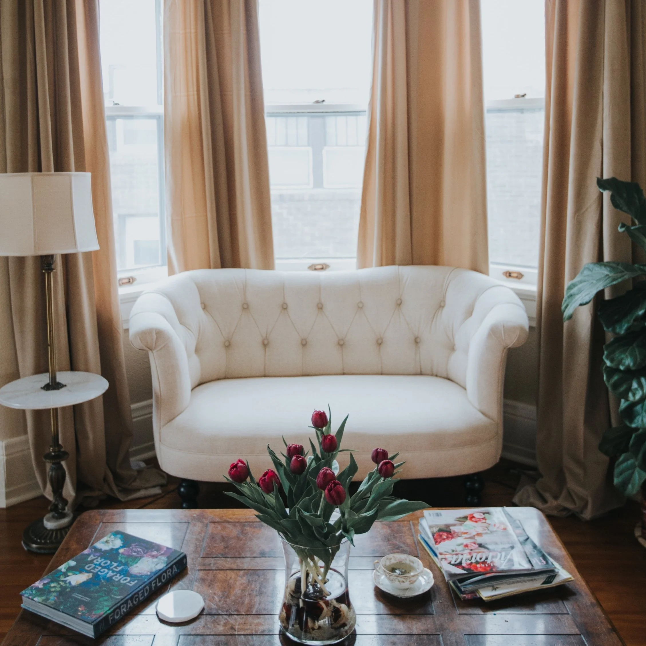 Historic sitting room with a white tufted settee, tall windows, layered side tables, and a floral arrangement.