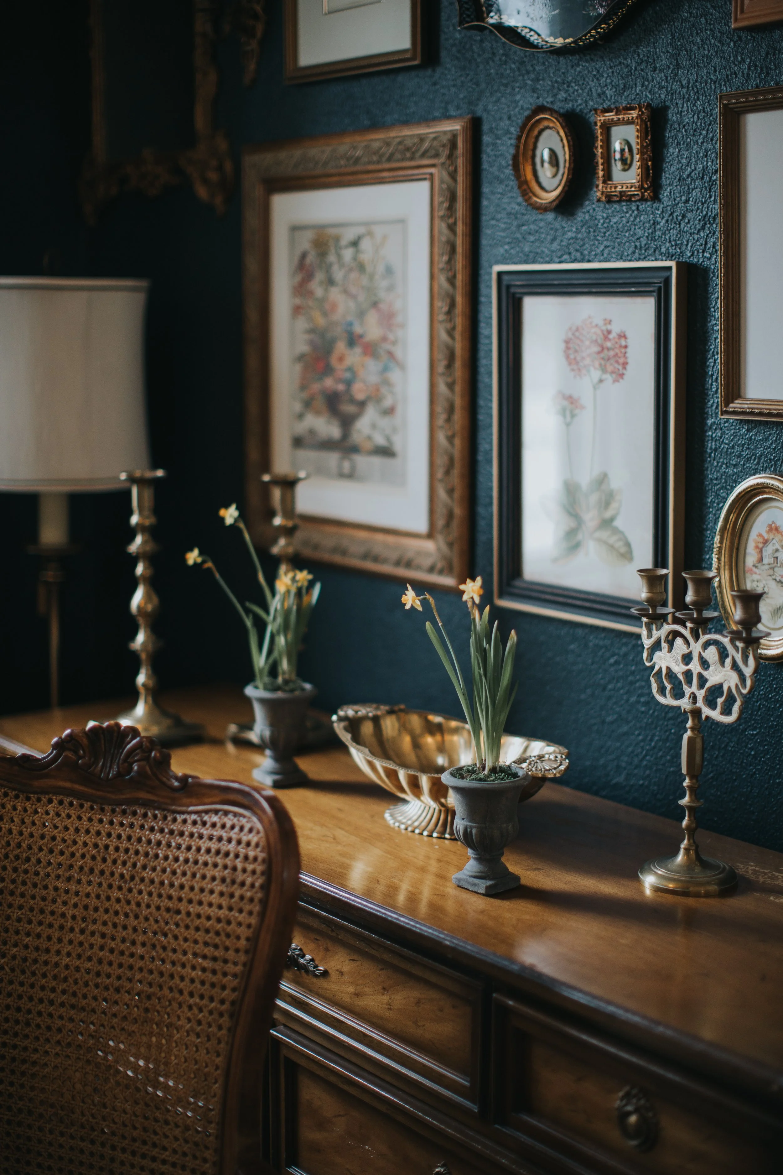 Antique wood sideboard styled with framed art, brass candlesticks, and potted bulbs in a dark blue dining room.