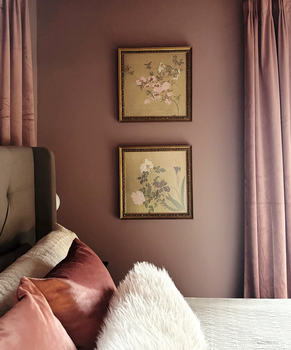 Mauve bedroom with matching curtains, framed botanical prints, rust-colored pillows, and a white faux-fur throw.