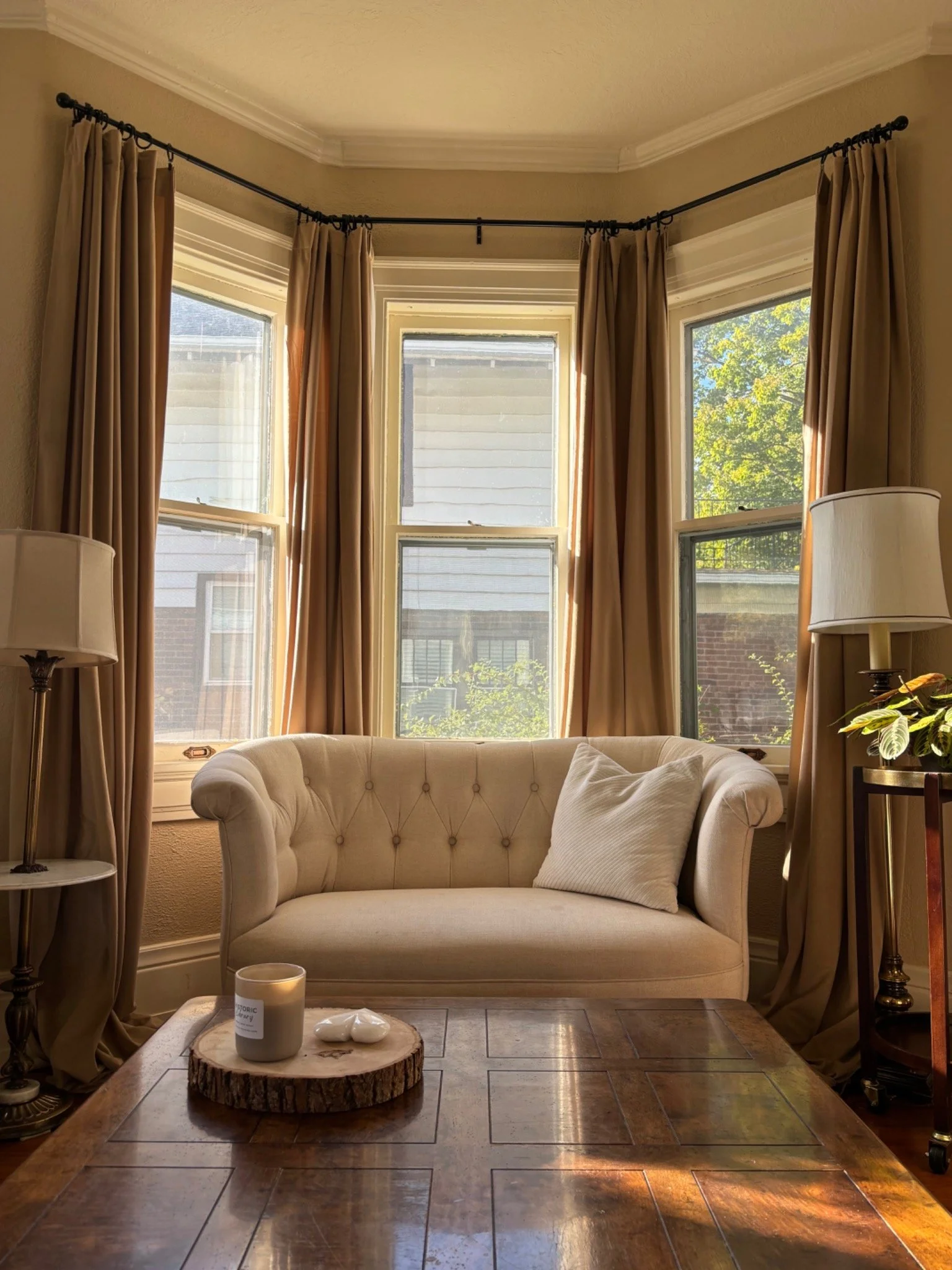 Sunlit traditional living room designed by Summer Glen Design with a cream tufted loveseat, floor-length neutral drapery, antique wood coffee table, and warm natural light.