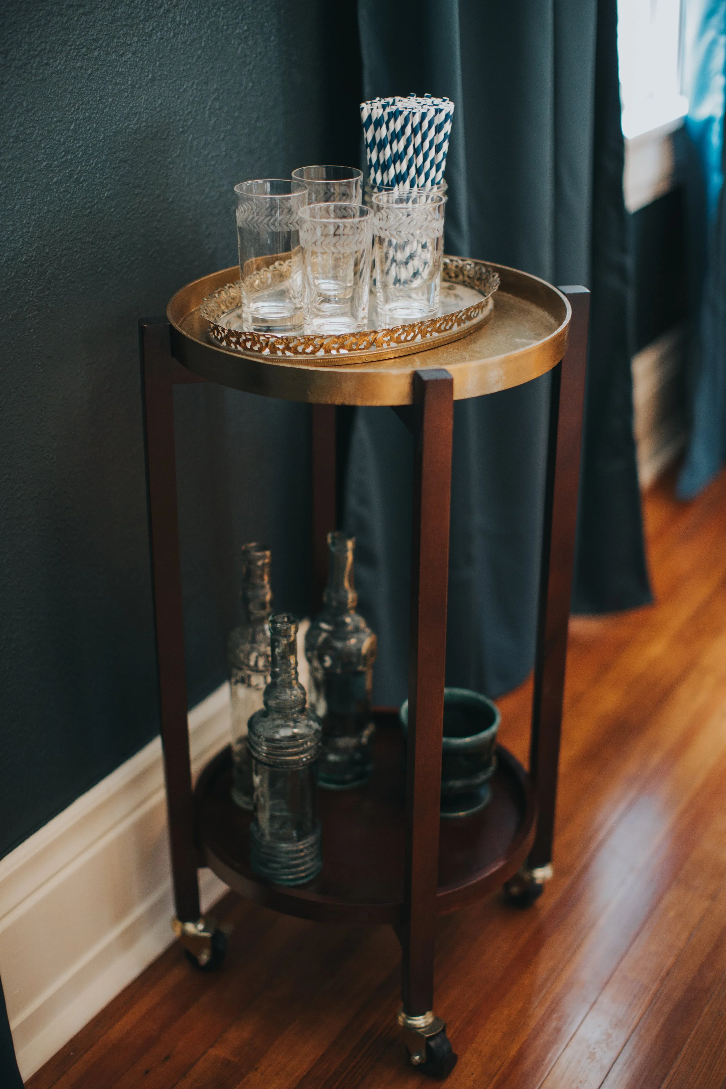 Antique round wood bar cart with crystal glassware and bottles against a dark blue wall and velvet curtains.