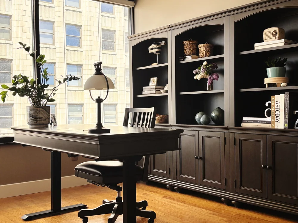 Home office with wall-to-wall dark wood cabinetry, open shelving, and a writing desk positioned in front of large windows.