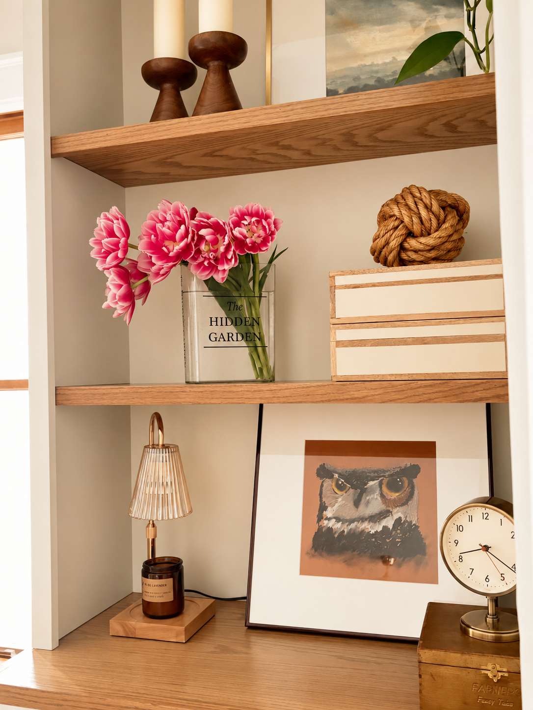 Built-in wood shelves styled with pink flowers, stacked books, framed art, sculptural objects, and a vintage clock.