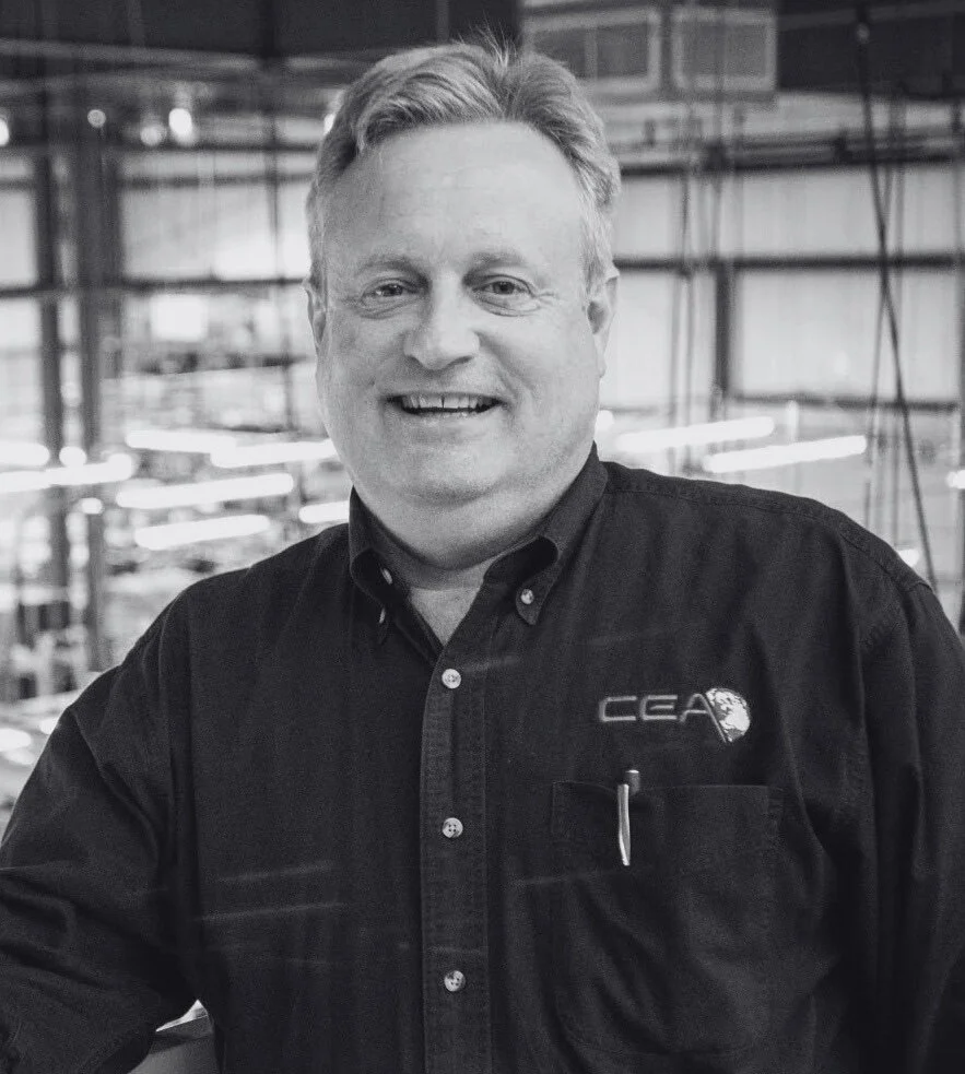 Man with short gray hair and wearing a dark button up shirt smiles at camera while standing in front of a faded industrial background