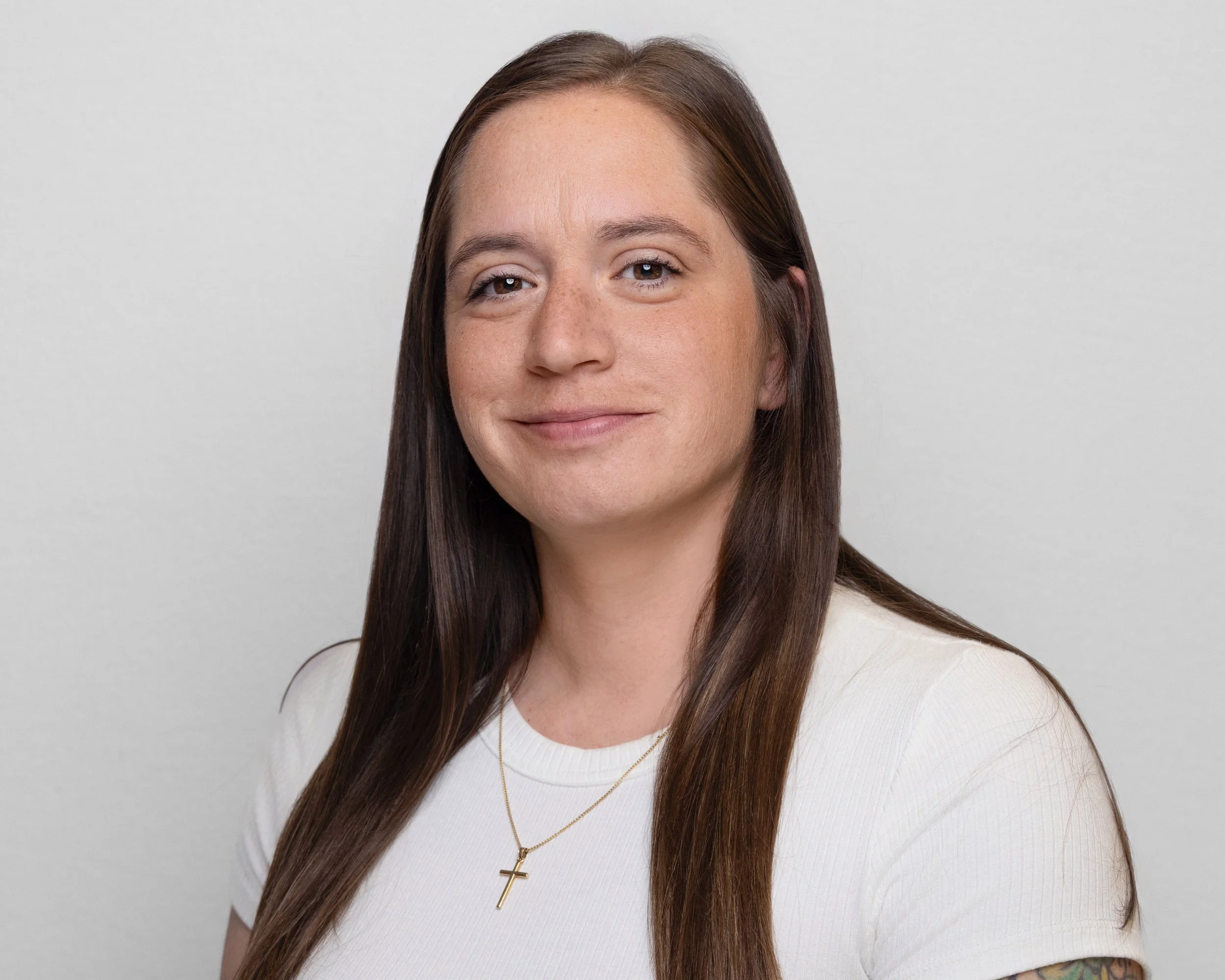 A woman with long brown hair wearing a white top and a gold cross necklace smiling at the camera against a plain light gray background.