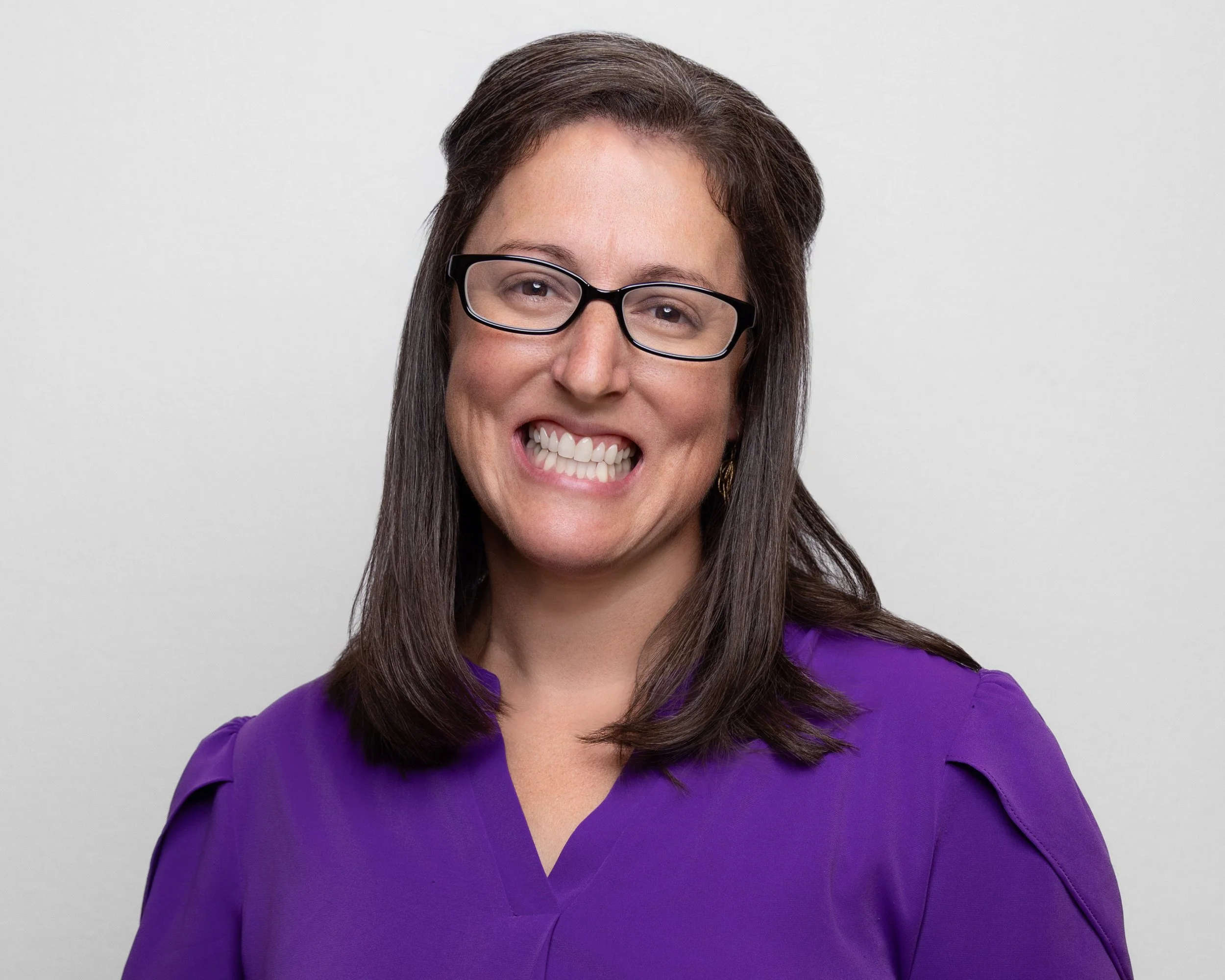 A woman with brown hair, glasses, and a purple top smiling with her teeth showing against a plain light gray background.