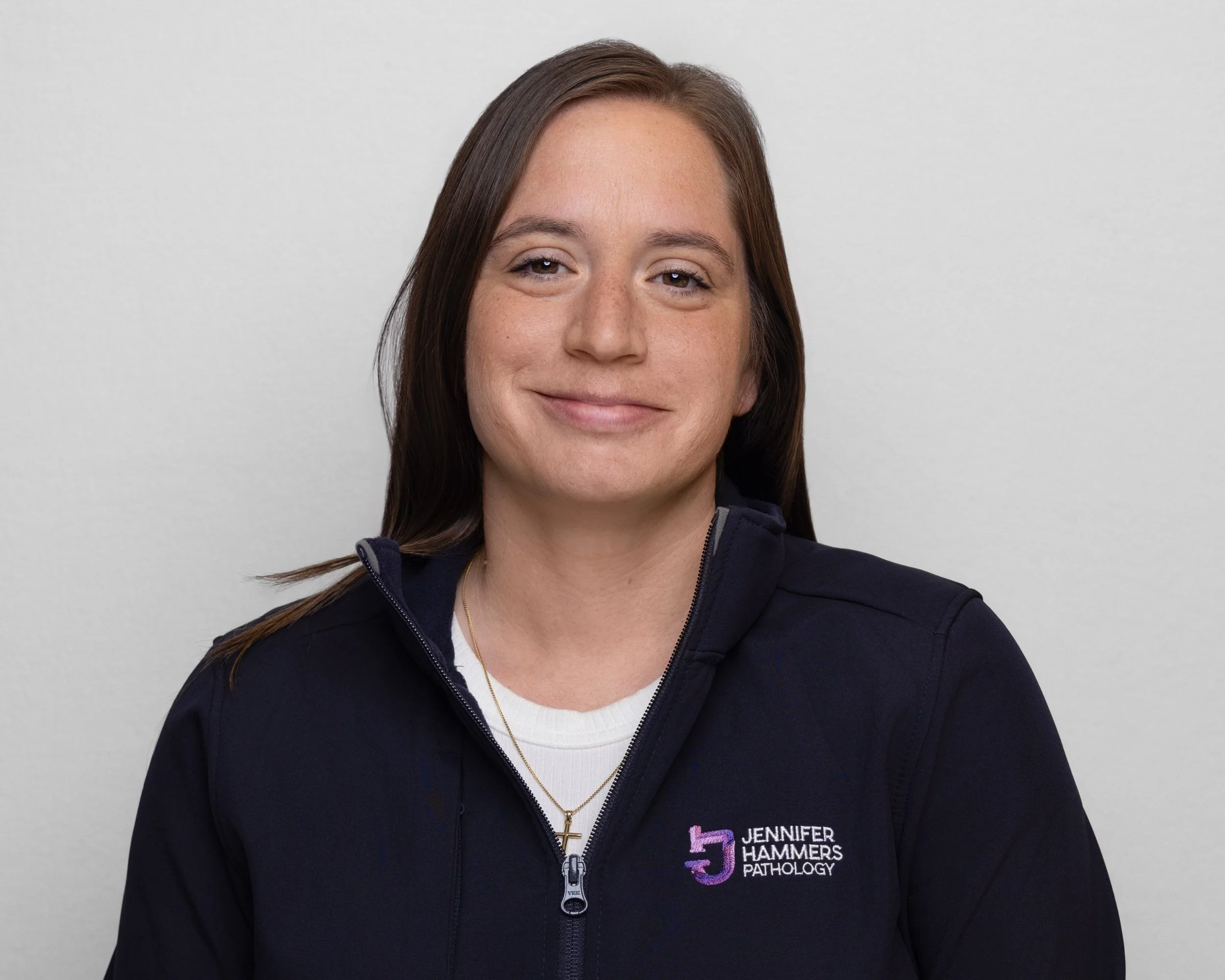 Woman with long brown hair smiling, wearing a navy blue jacket with a 'Jennifer Hammers Pathology' logo, standing against a plain light gray background.
