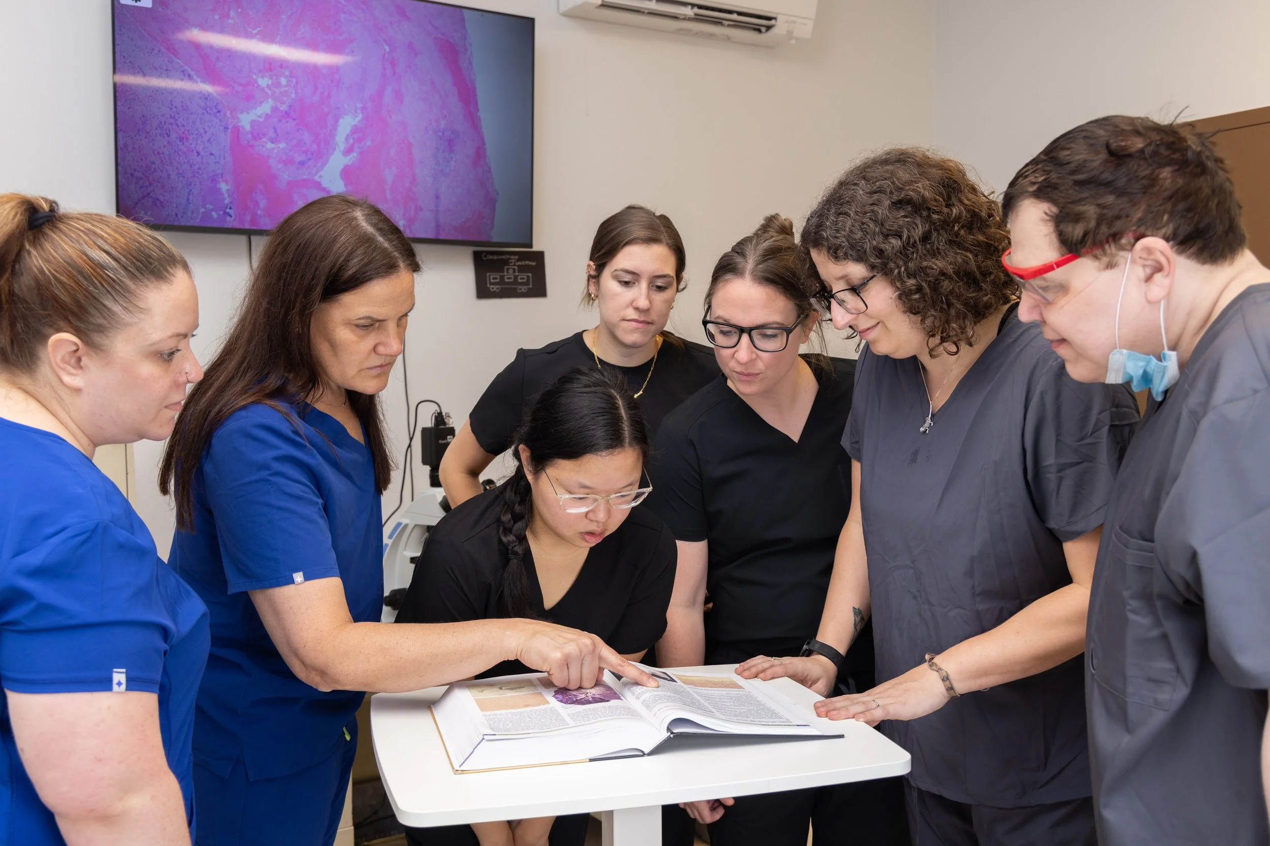 Group of seven medical professionals in scrubs and glasses gathered around a table looking at a large medical book, with a monitor displaying a pink and purple medical image in the background.