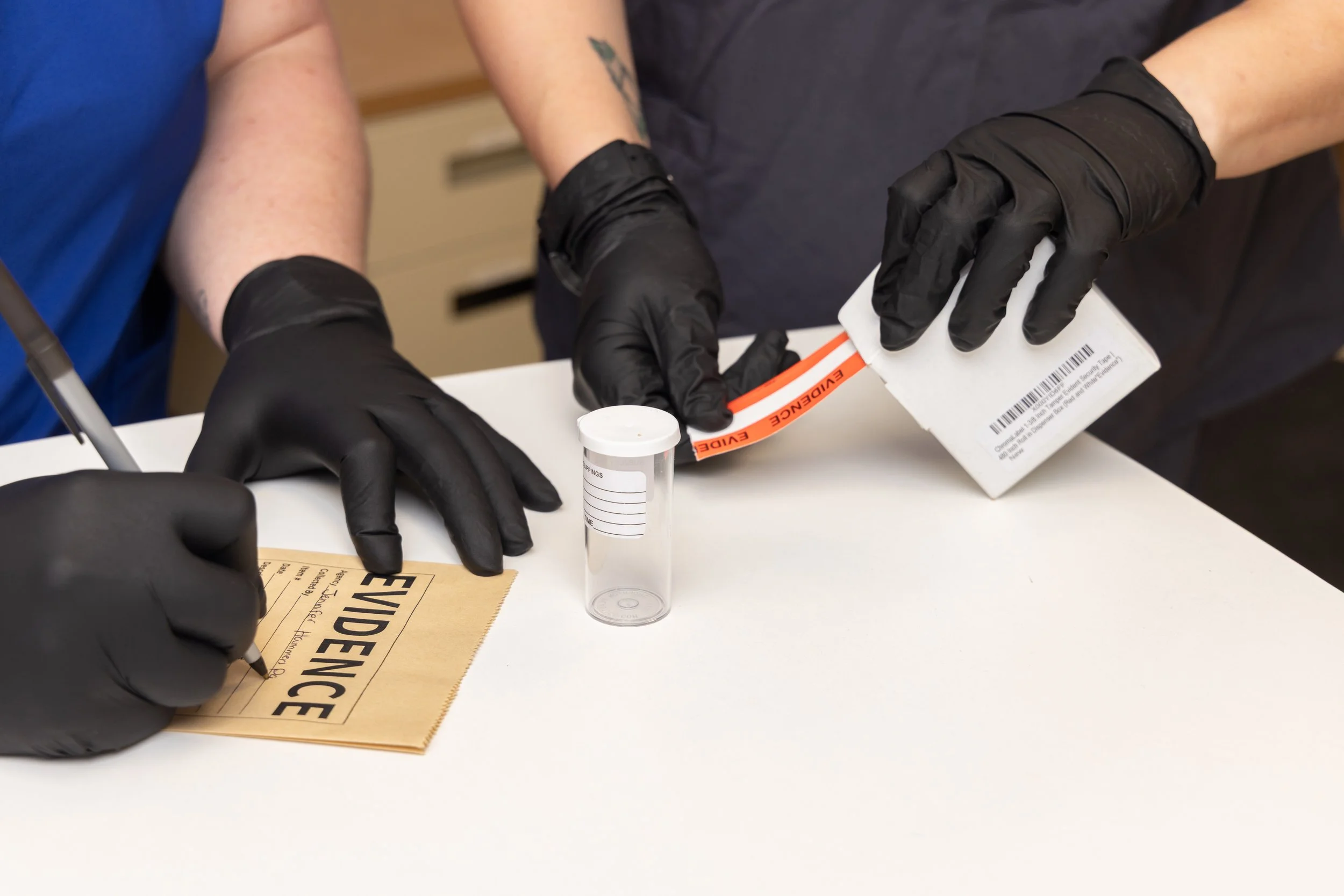 Two individuals wearing black gloves are handling a medical specimen label and a sample container on a white table, with a person signing a document labeled 'EVIDENCE' in a clinical setting.