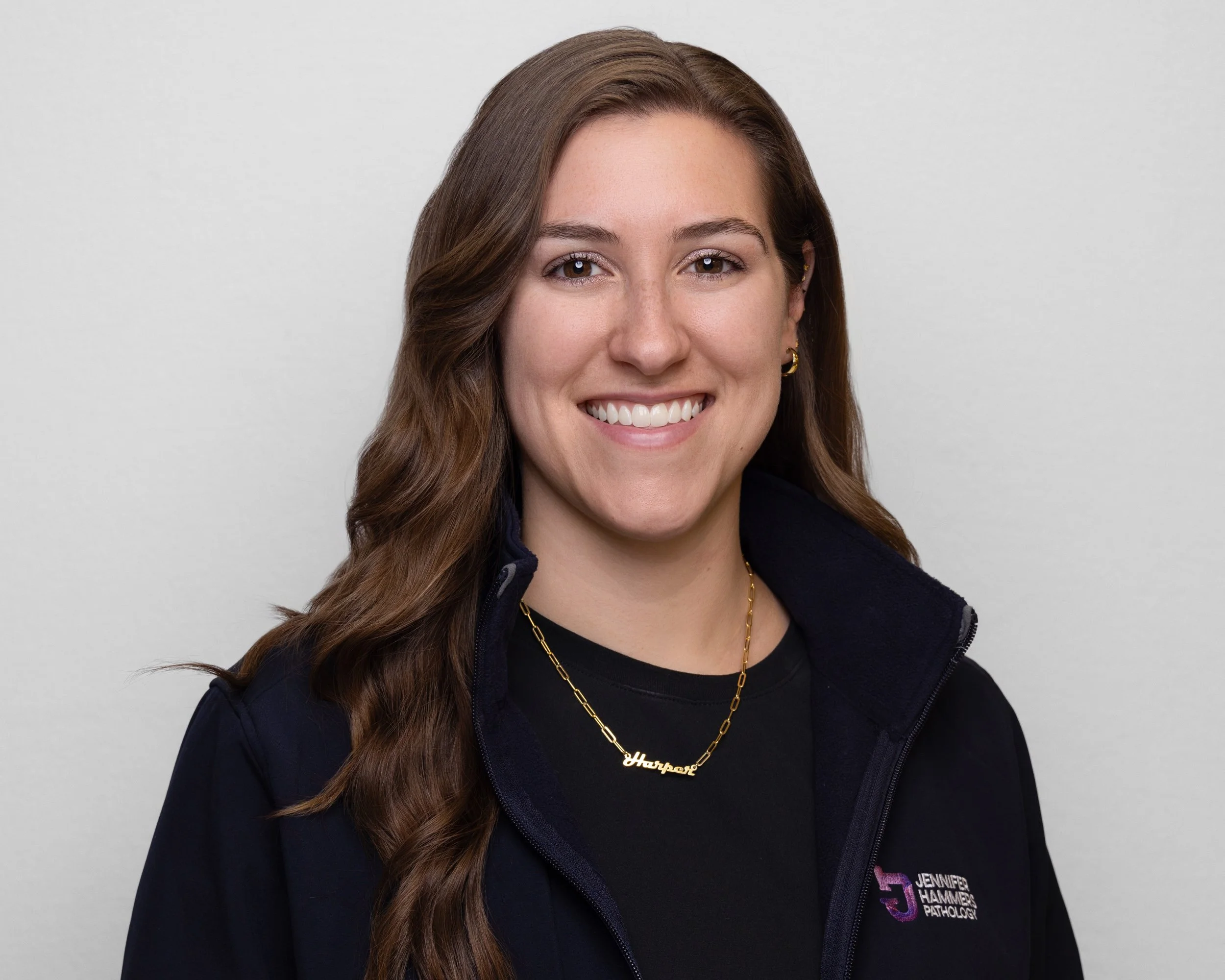 A woman with long wavy brown hair, smiling, wearing a black shirt, a navy jacket with a logo, and a gold necklace with a nameplate that says 'Harpak'.