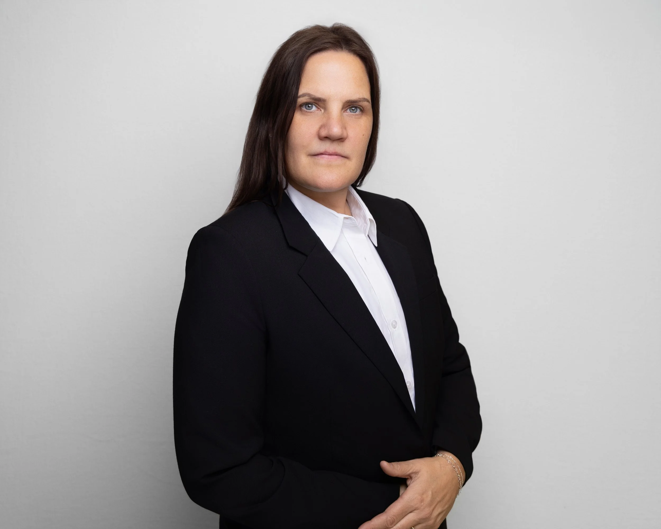A woman with long brown hair wearing a black blazer and white dress shirt standing against a plain light-colored background.