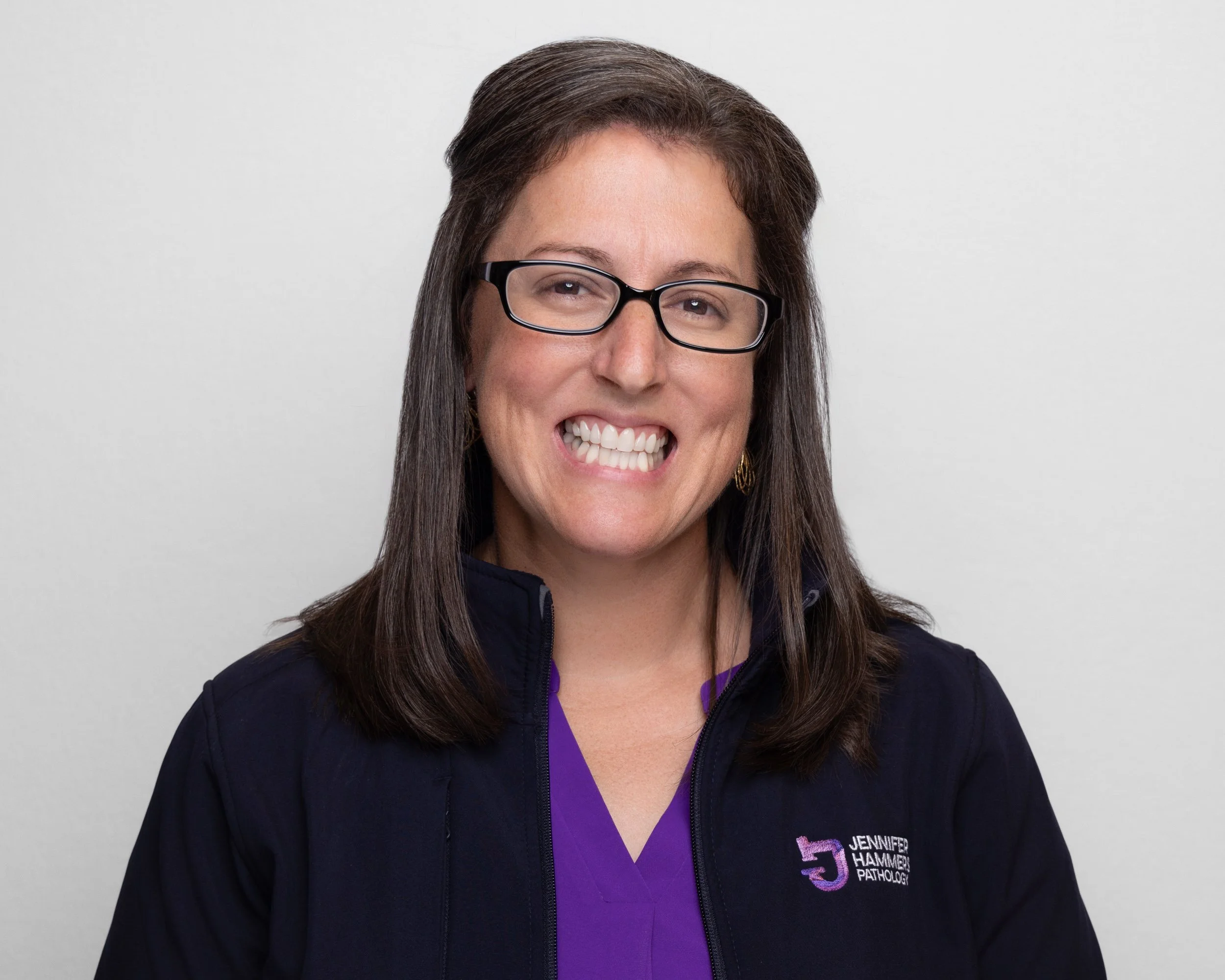 Portrait of a woman with shoulder-length brown hair, wearing glasses, a purple top, and a dark jacket with an embroidered logo that reads 'Jennifer Hammer Pathology,' smiling at the camera against a plain white background.