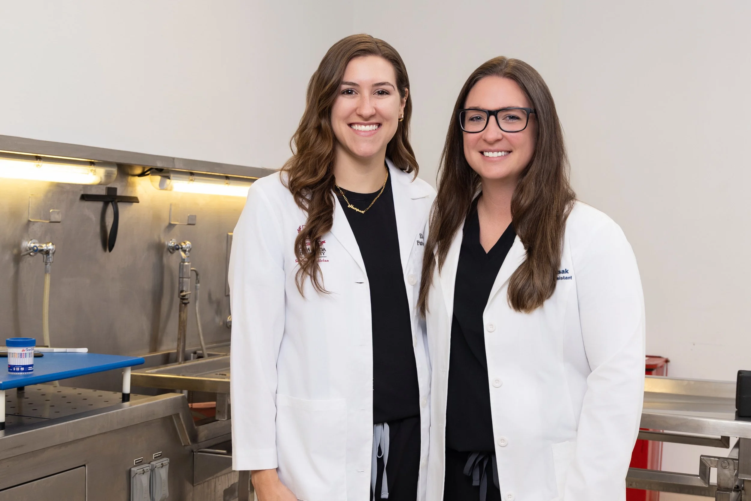 Two women in white lab coats standing in a laboratory, smiling at the camera.