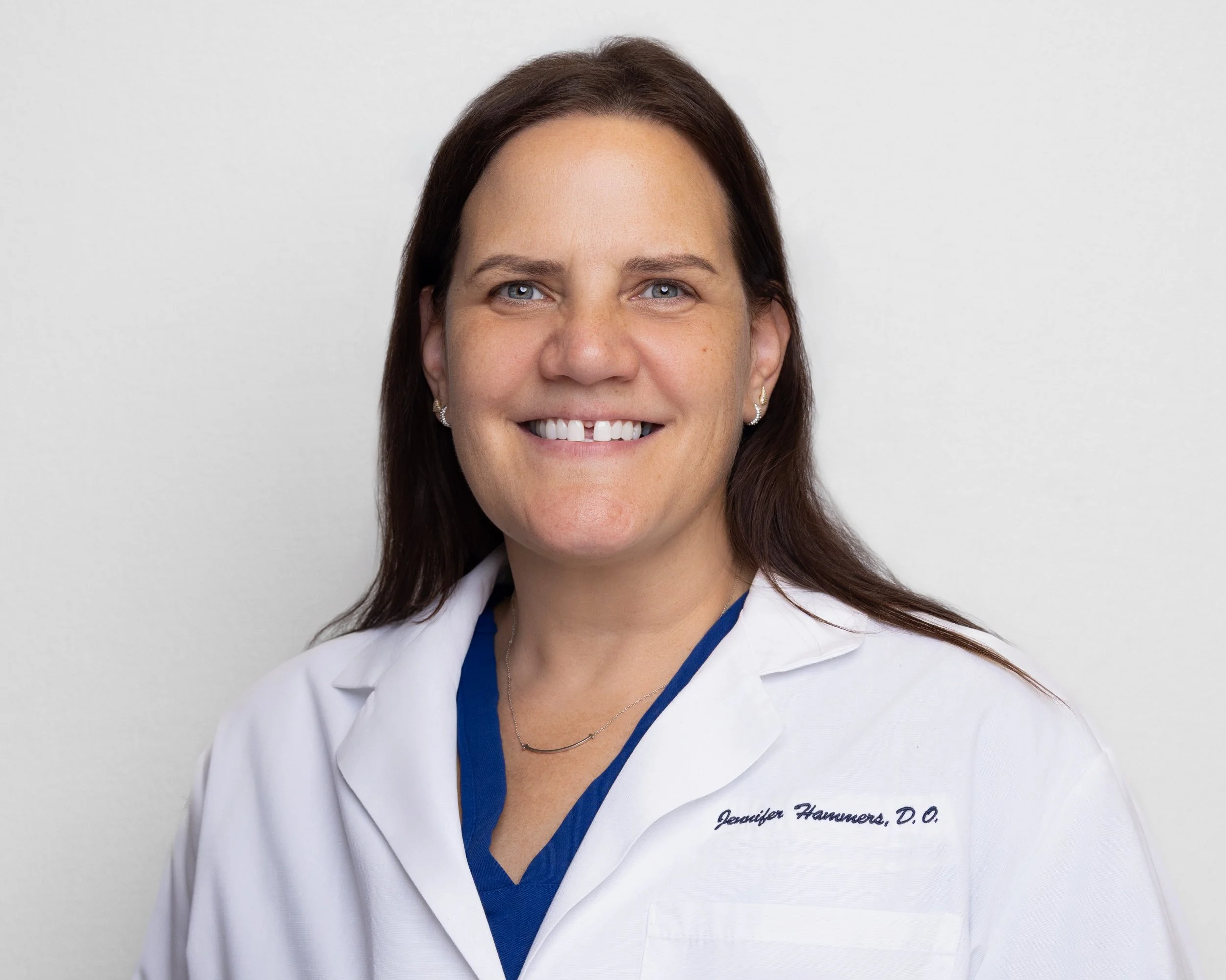 A smiling woman in a white medical coat with "Jennifer Hammers, D.O." embroidered on it, standing against a plain white background.