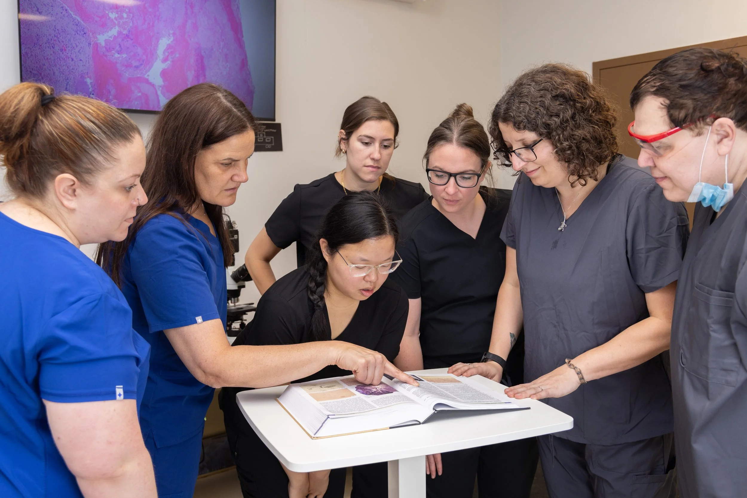 Group of medical professionals gathered around a table, looking at a large open book and discussing something, with some wearing scrubs and one person pointing at the book.