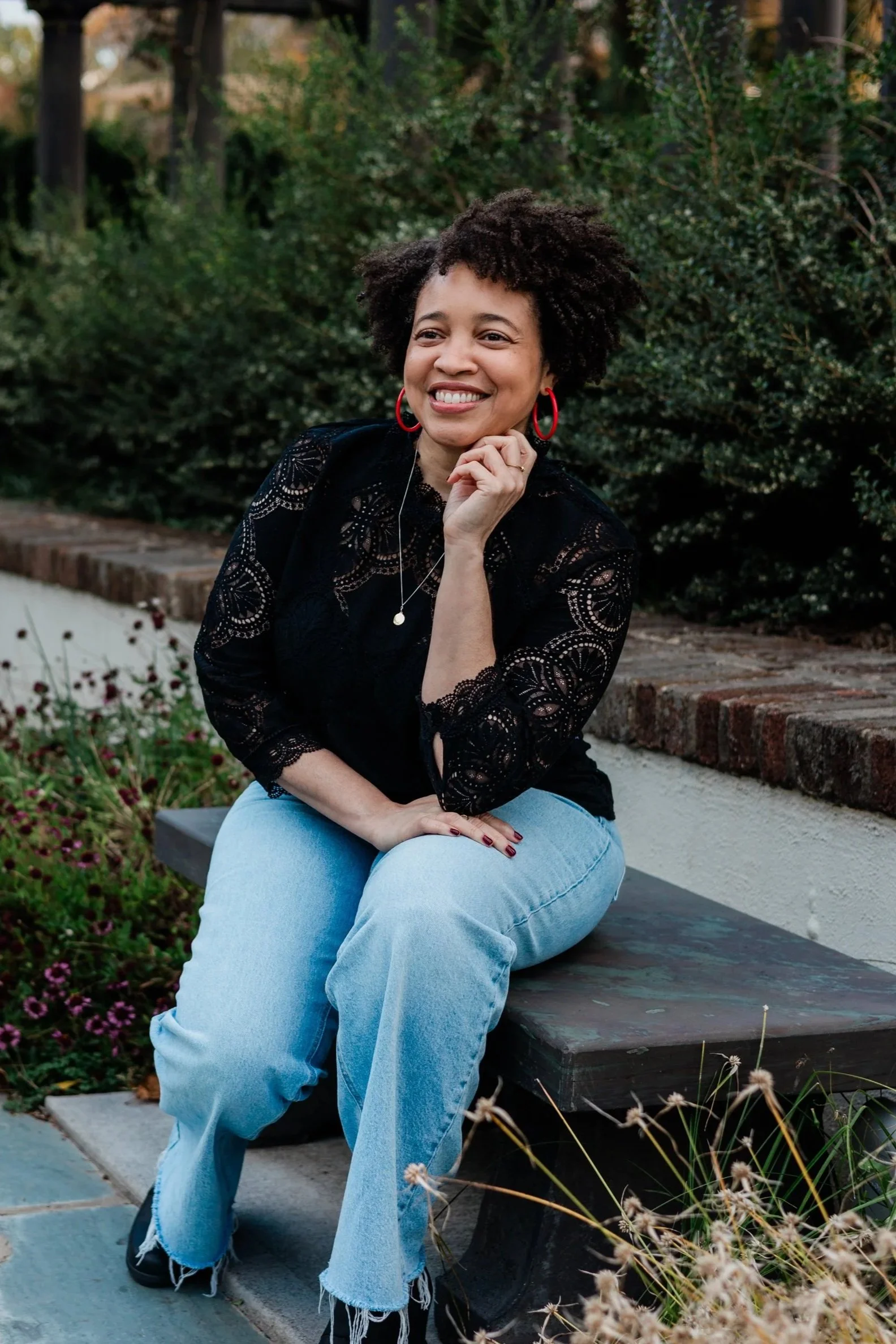 A woman with curly hair sits on a bench outdoors, smiling, wearing a black lace top, light blue jeans, red hoop earrings, and a necklace, surrounded by greenery and flowers.