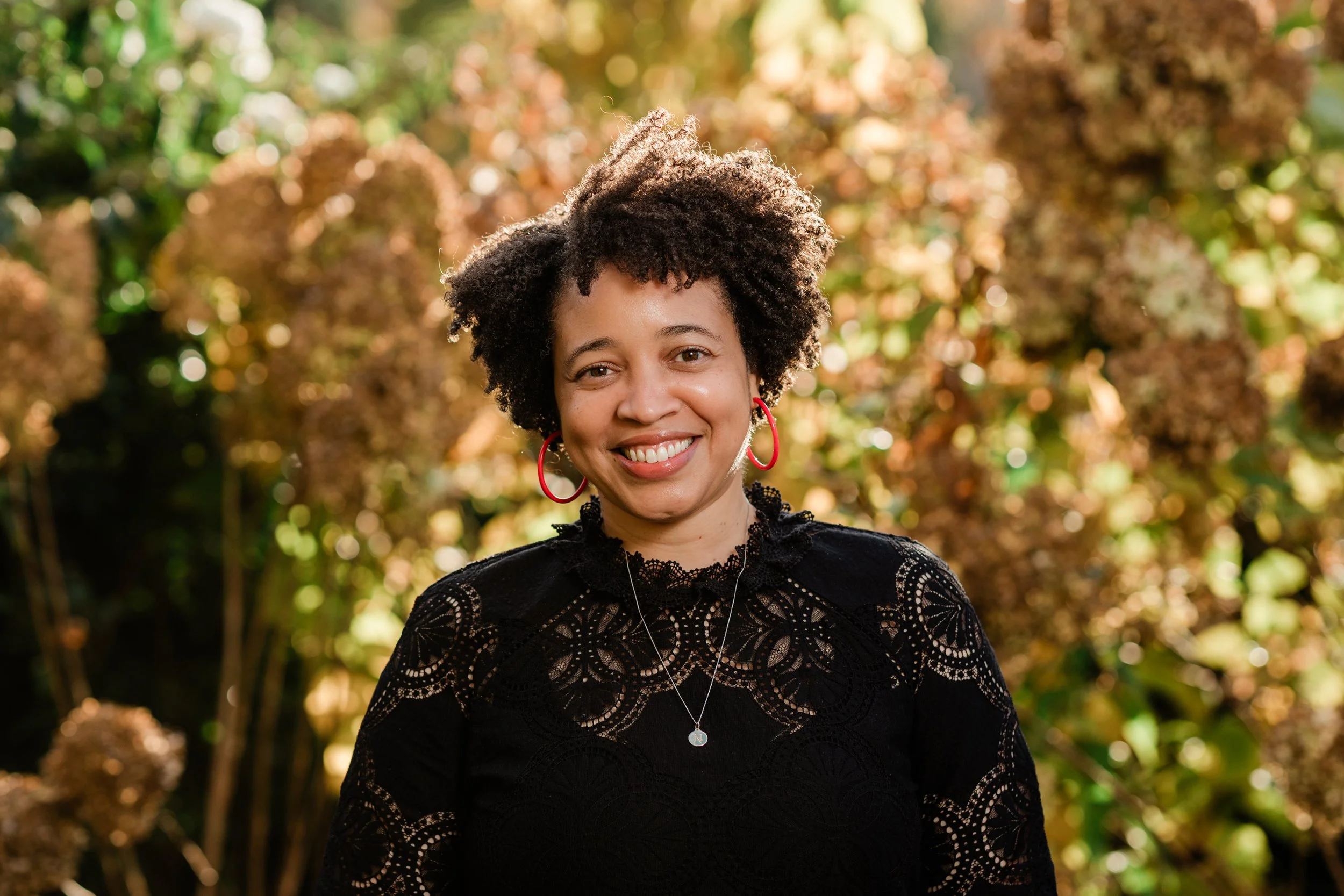 A smiling woman with curly hair and red hoop earrings posing outdoors during fall, wearing a black lace dress with a necklace.