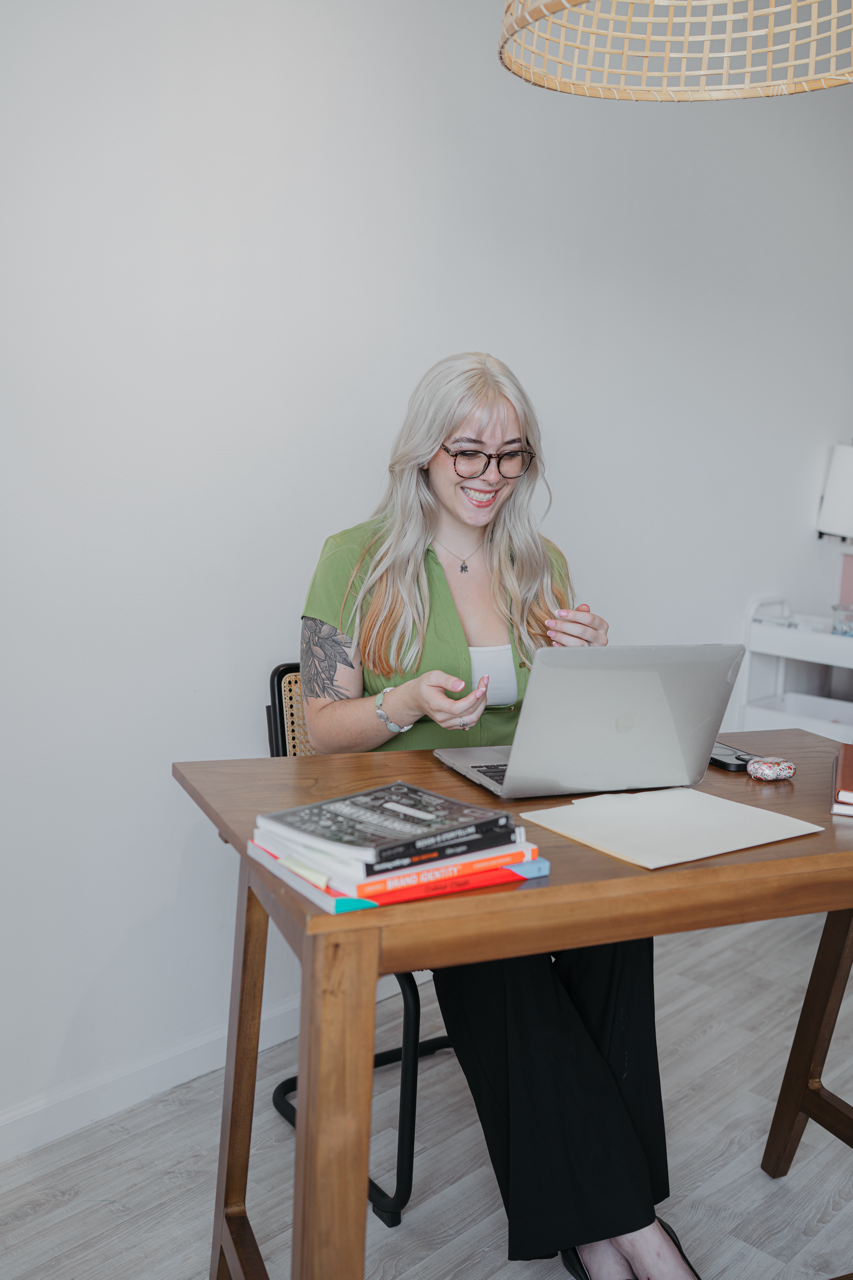 A smiling woman with long blonde hair, glasses, and tattoos on her arm sitting at a wooden desk with a laptop, books, and notebooks, in a bright room.
