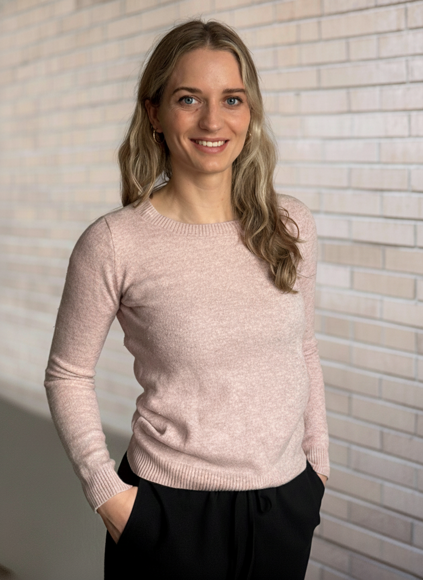 Junge Frau mit blauen Augen und blonden, gewellten Haaren vor einer Backsteinwand, trägt ein rosa Pullover und schwarze Hosen, lächelt.