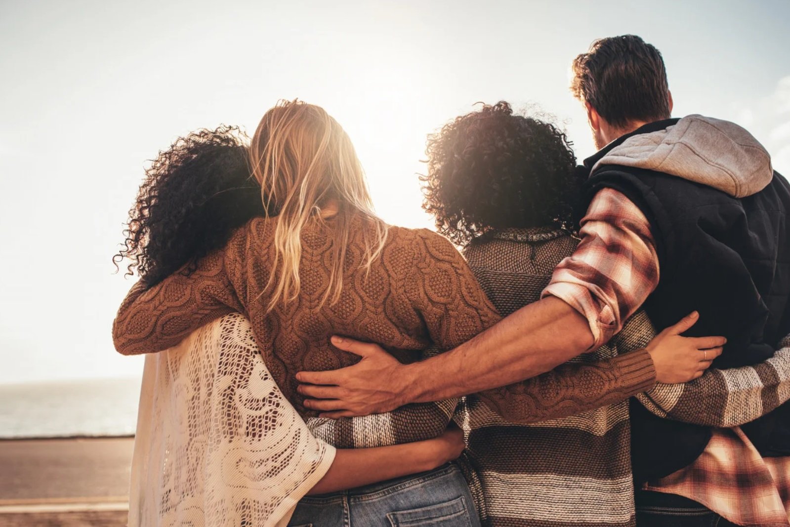Vier Menschen, die sich an einem sonnigen Tag an einem Strand umarmen und zusammenstehen, mit Blick auf das Meer im Hintergrund.