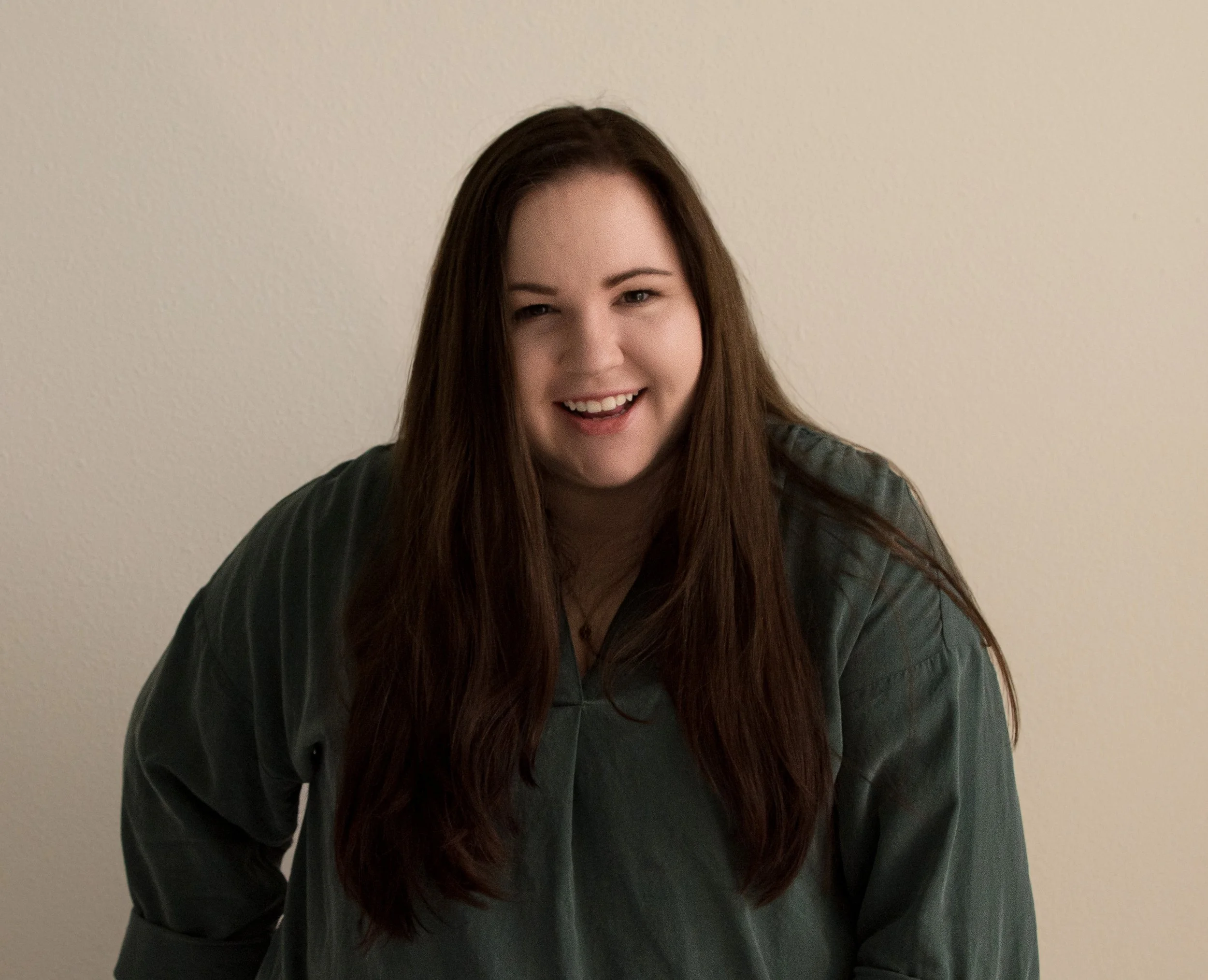 A smiling woman with long brown hair wearing a loose gray shirt, standing against a plain beige wall.