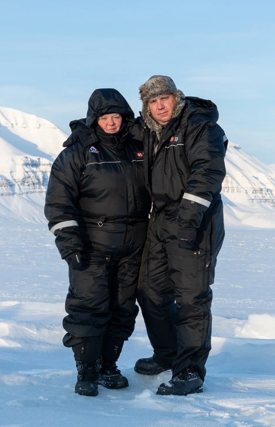 Two people standing together in snowy mountainous environment, dressed in heavy black winter gear with hoods and fur-lined hats, suggesting they're in a cold, Arctic-like setting.