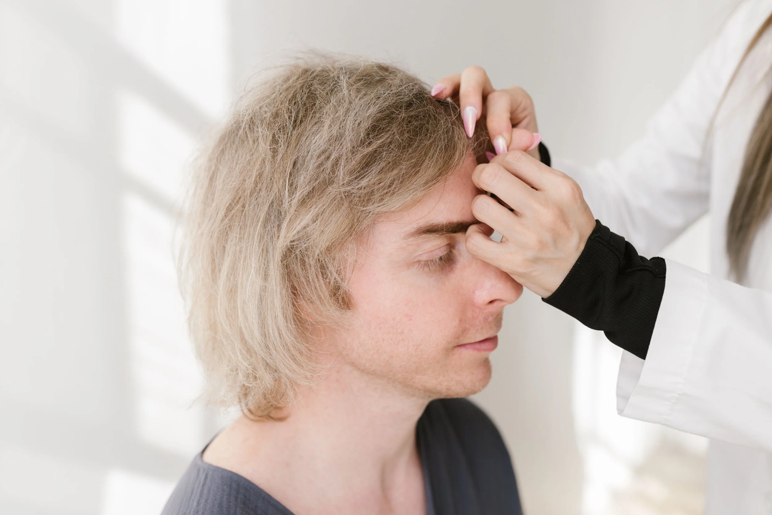 Close-up of a person with light-colored hair and closed eyes receiving a forehead massage from a person wearing a white coat in a clinical setting.