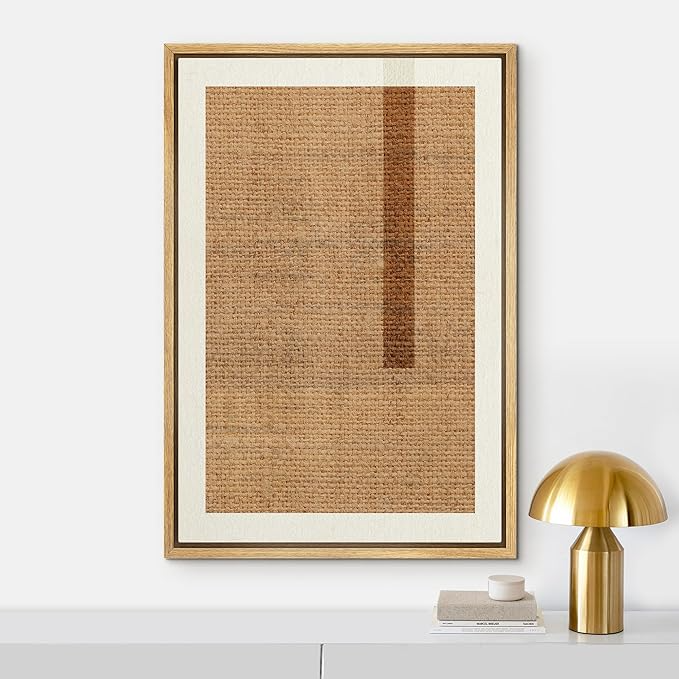 Framed modern abstract artwork with textured brown and dark brown rectangular shapes hanging on a white wall, with a gold table lamp and a small stack of books on a white surface below.