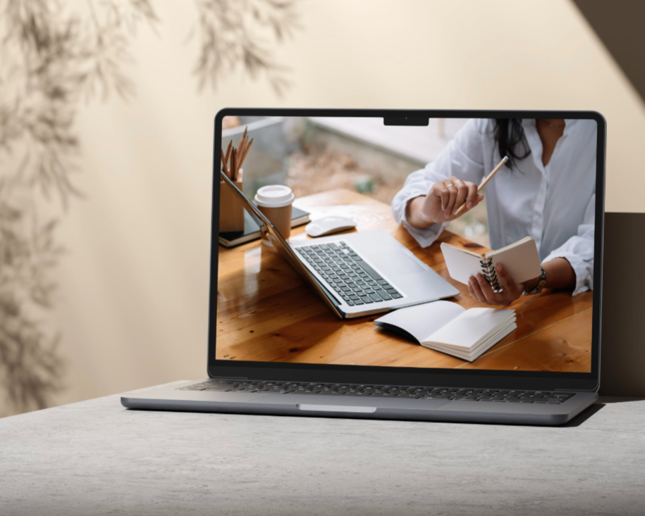 A person on a video call, sitting at a desk with a laptop, notebooks, pen, and coffee cup, with a blurred indoor background.