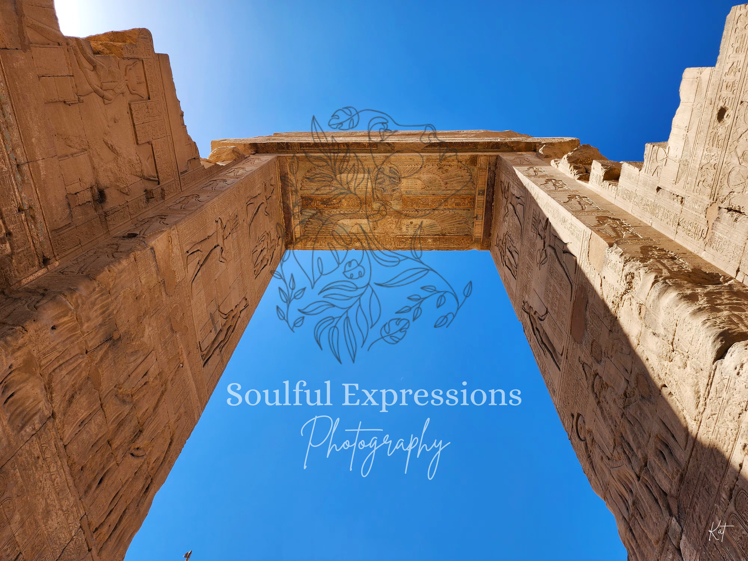 Looking up at the entryway of the Dendera Temple Complex in Egypt, with columns with hieroglyphics, under a bright blue sky with a faint outline of a bird and clouds.
