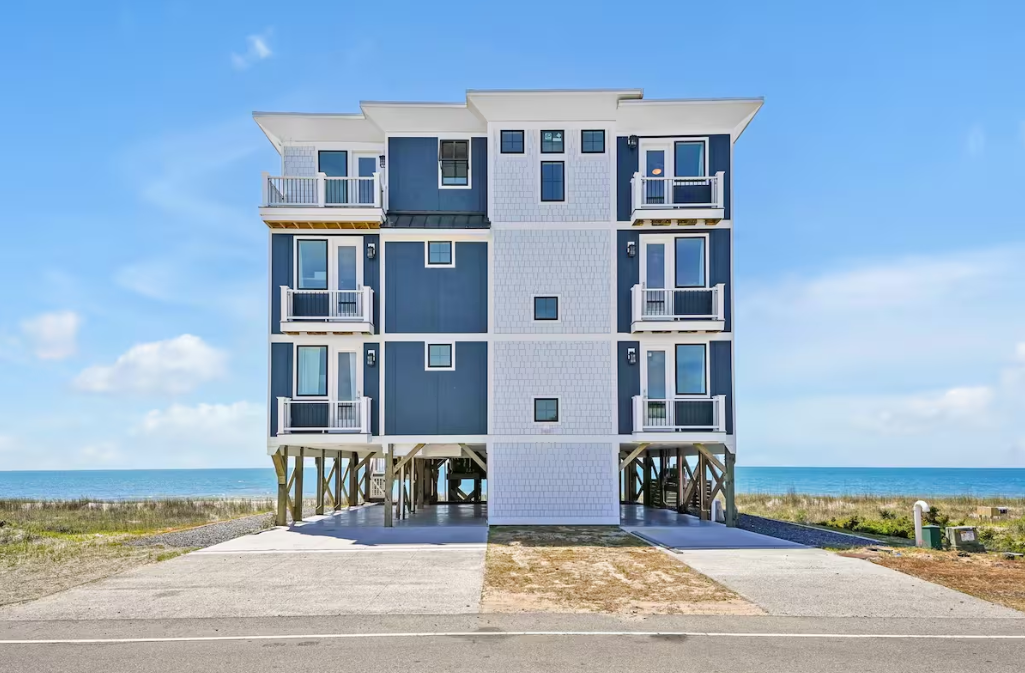 A multi-story beach house elevated on stilts facing the ocean with a driveway and a clear blue sky.