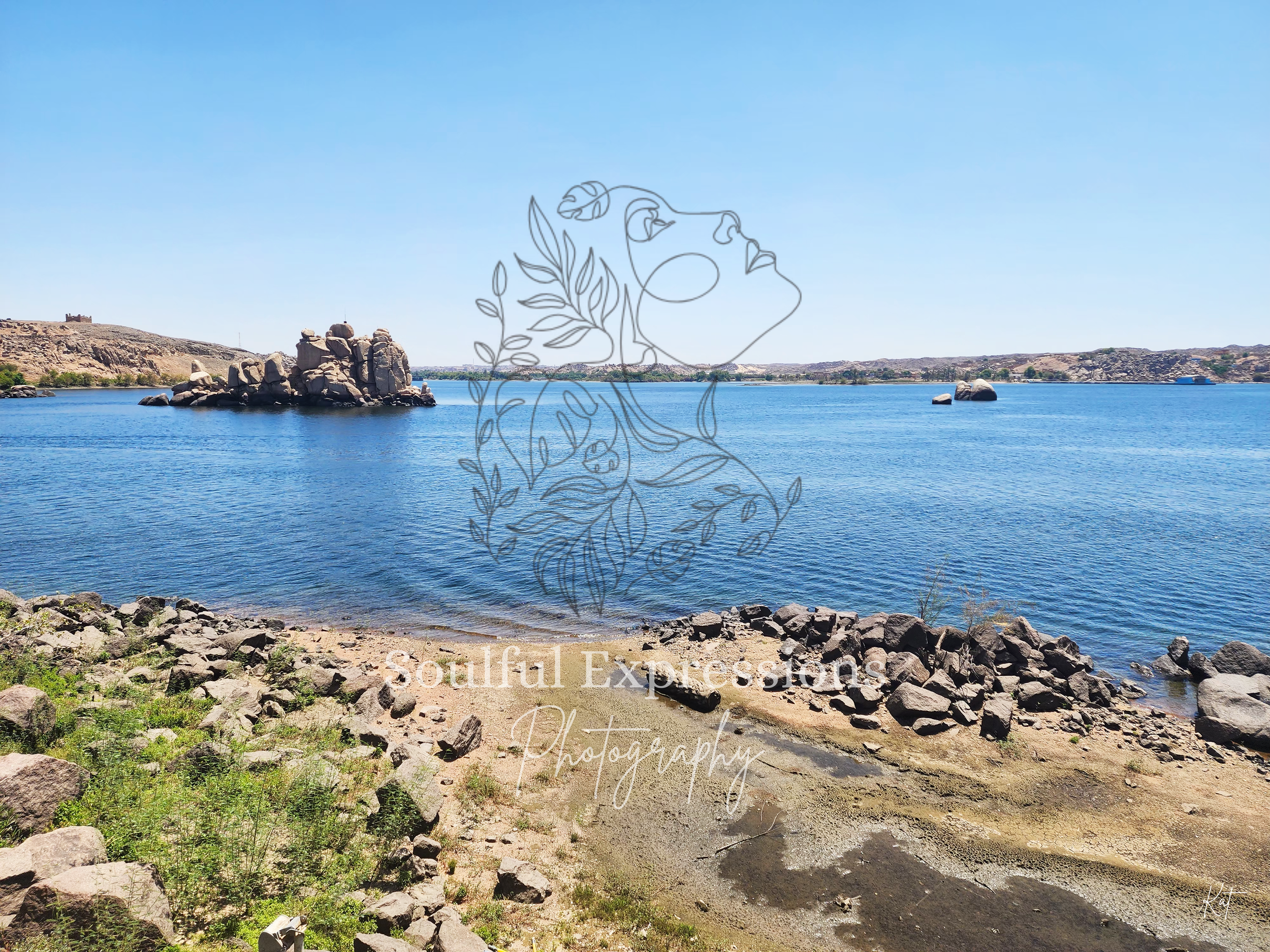 A scenic view the Nile River outside of Isis Temple in Egypt with rocky shoreline, clear blue water, and distant hills under a bright blue sky.