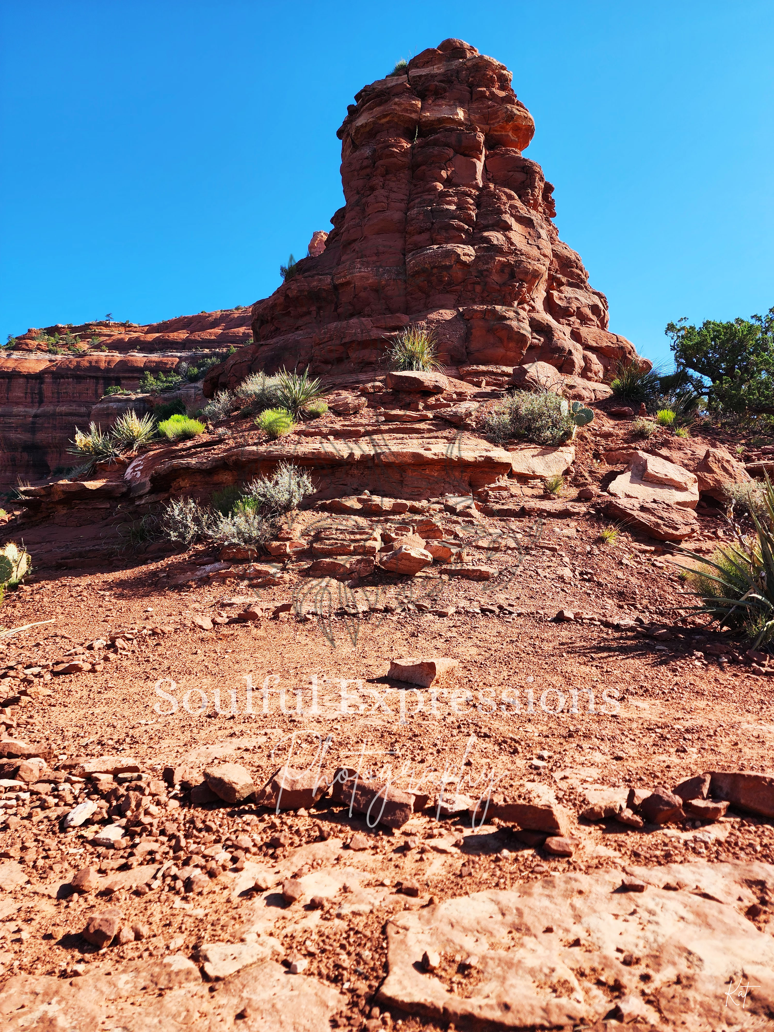 A large red rock formation in a desert landscape with sparse green vegetation and a clear blue sky in Sedona, Arizona.