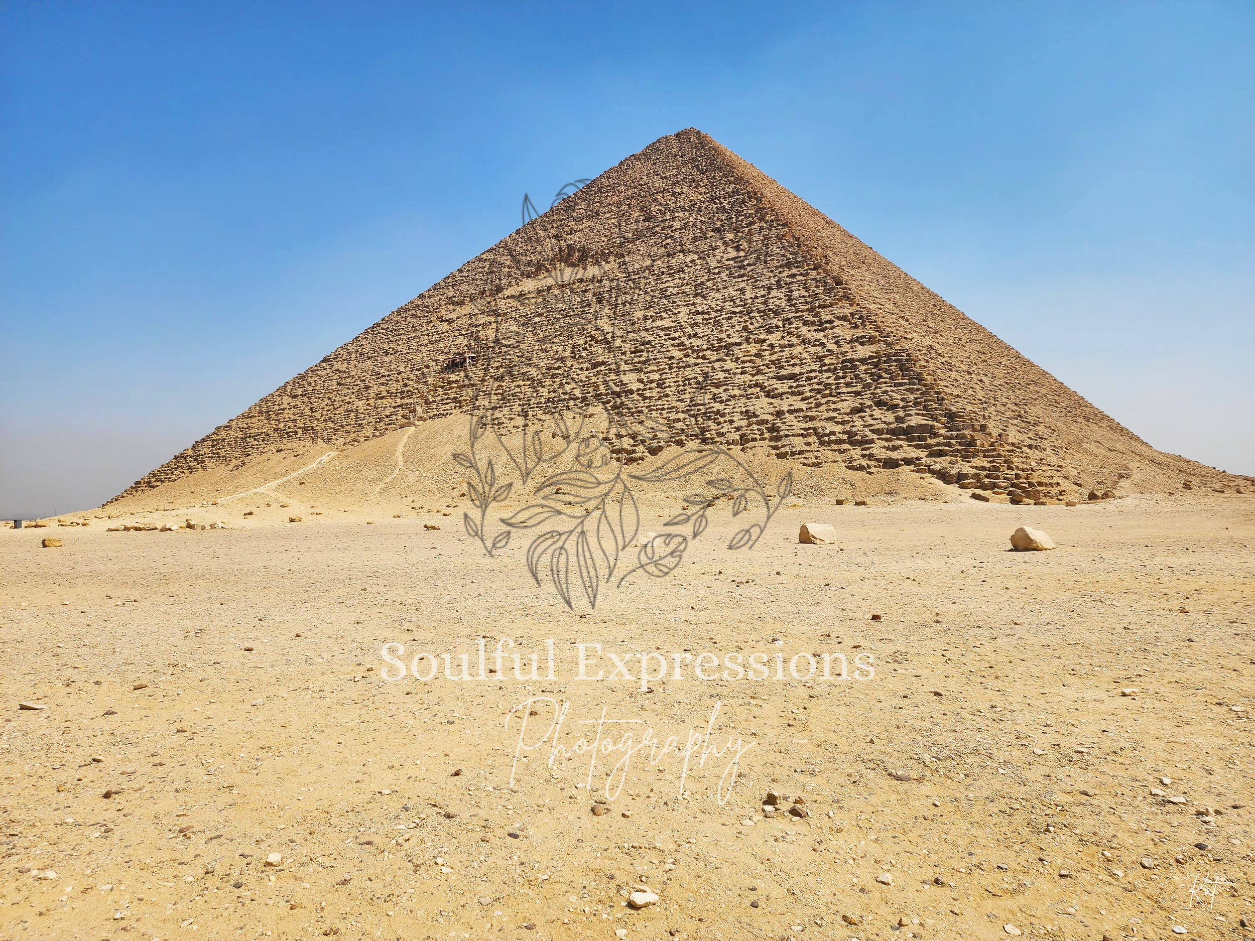 The Red Pyramid in Egypt in the desert under a clear blue sky with a few scattered rocks on the ground.