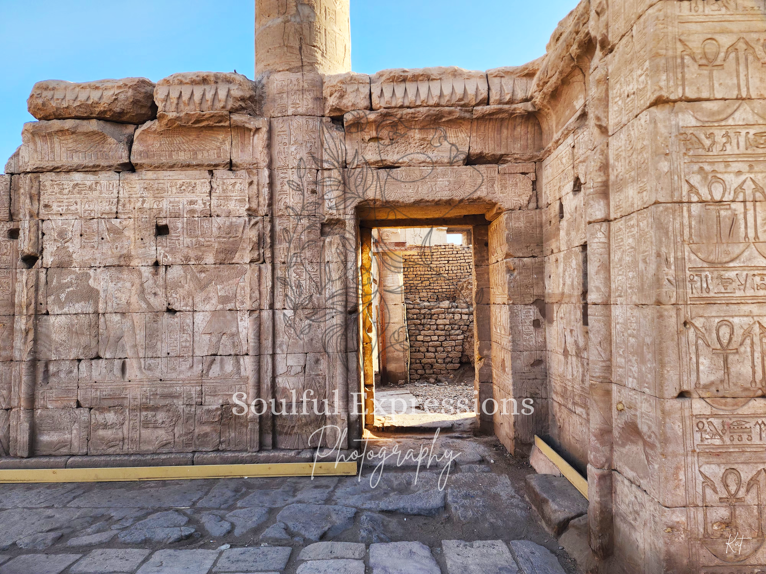 Ancient Egyptian stone wall inside of the Dendera Temple Complex in Egypt with hieroglyphics and carvings, featuring an archway leading to a brick wall in the background.