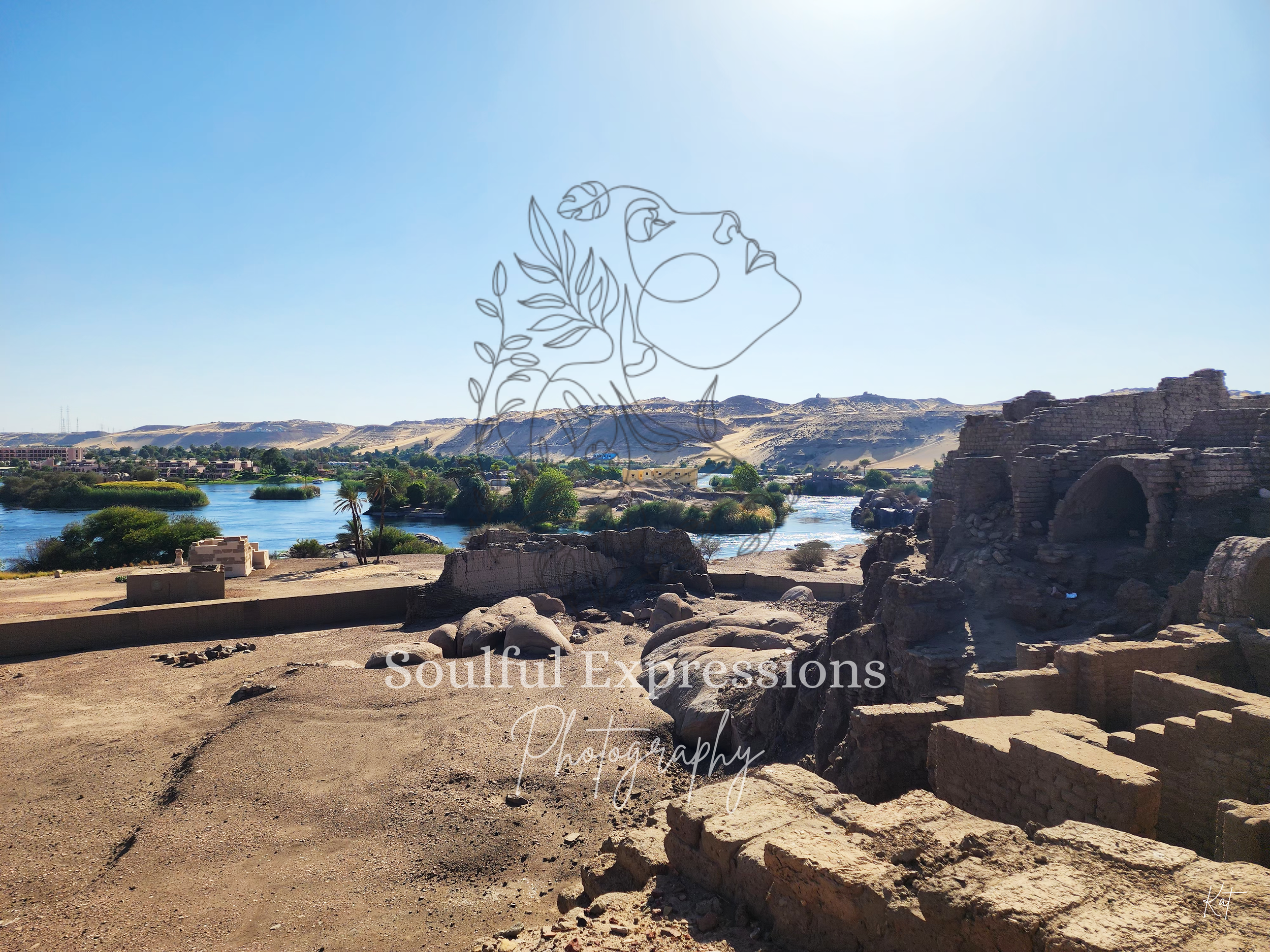 Ancient ruins with stone structures and arches, a river flowing in the background, desert hills, and sparse vegetation under a clear blue sky.
